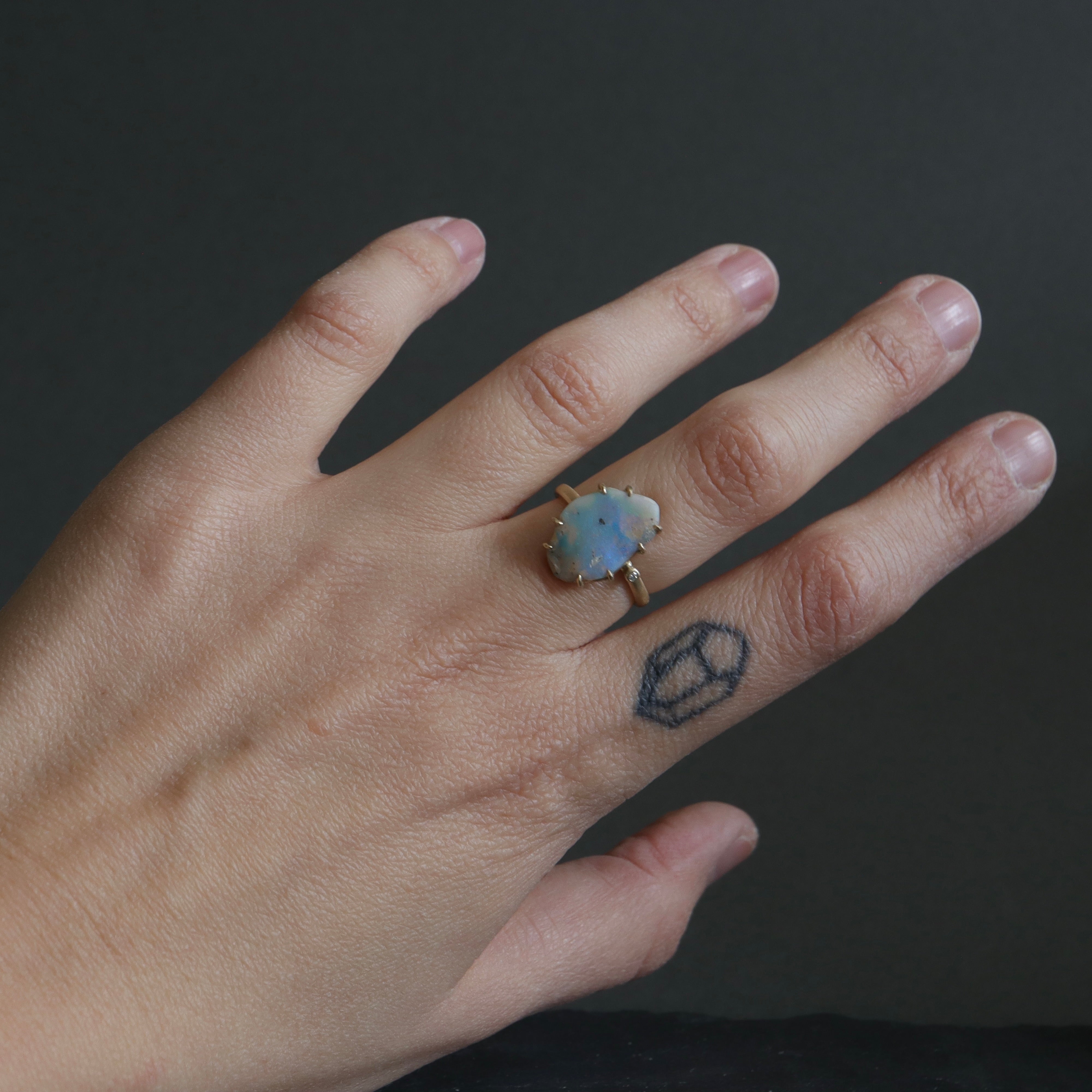 raw opal with rainbow colors is set within eight gold prongs on a gold band. A single diamond sits atop the band next to the opal, like a little friend. Shown on person.