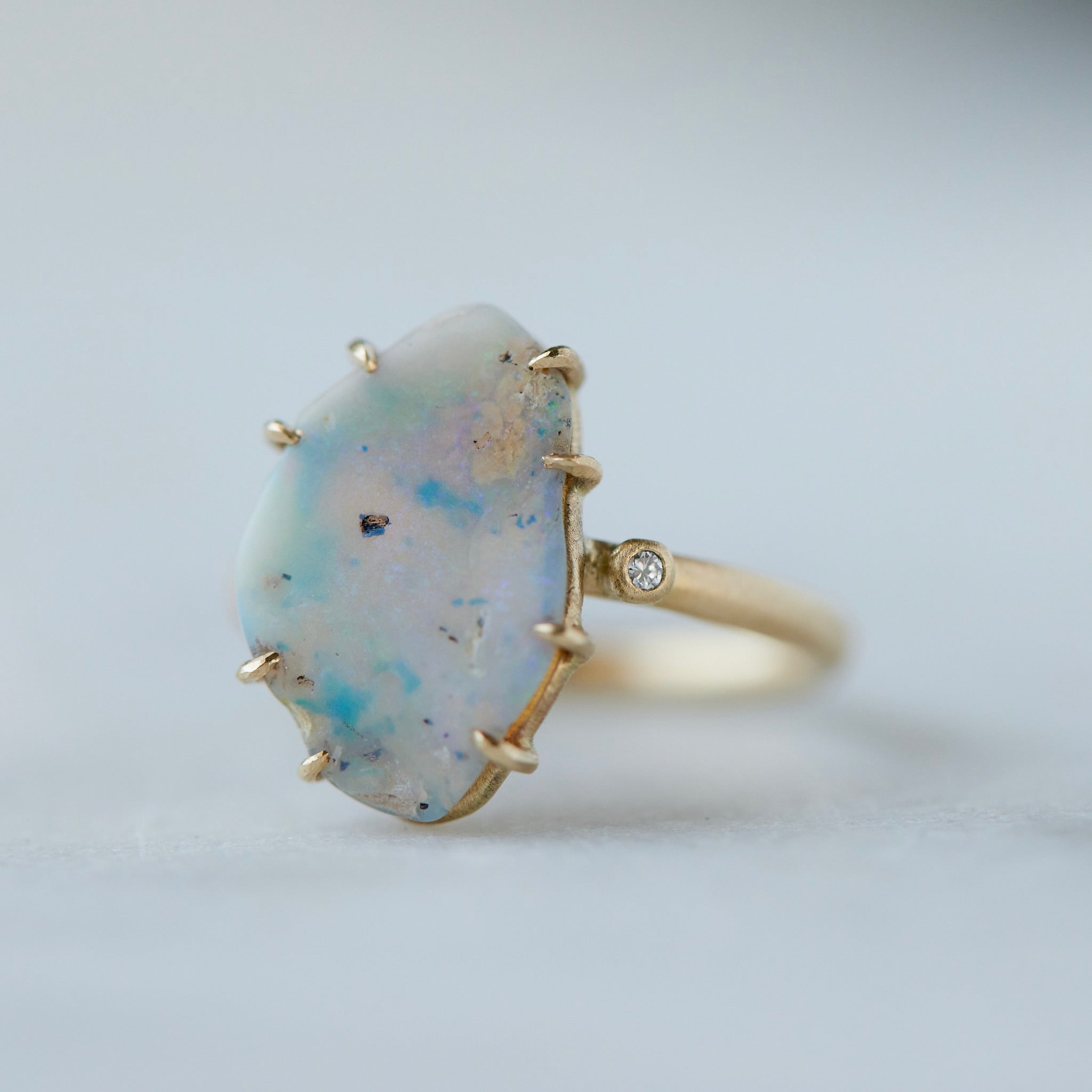 raw opal with rainbow colors is set within eight gold prongs on a gold band. A single diamond sits atop the band next to the opal, like a little friend.