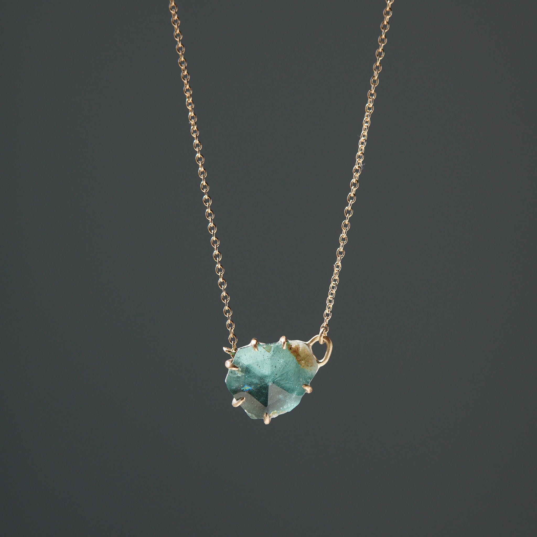 partially polished, partially raw blue-green tourmaline gemstone is set in gold prongs and has a gold chain attached to each side.