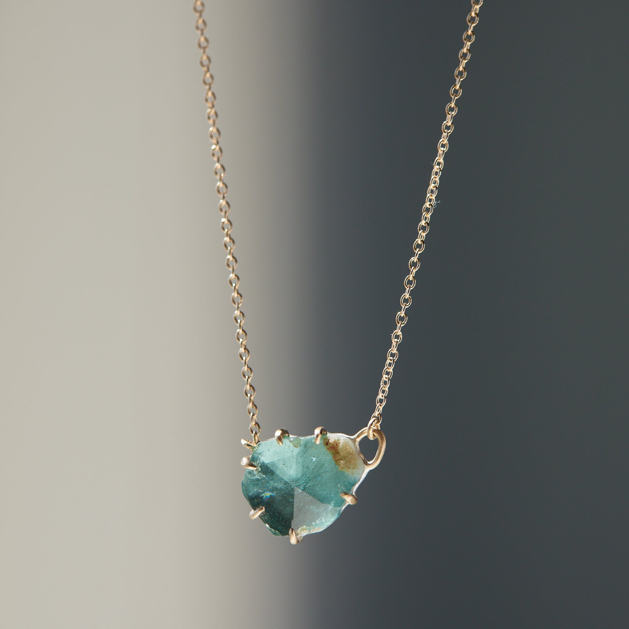 partially polished, partially raw blue-green tourmaline gemstone is set in gold prongs and has a gold chain attached to each side.