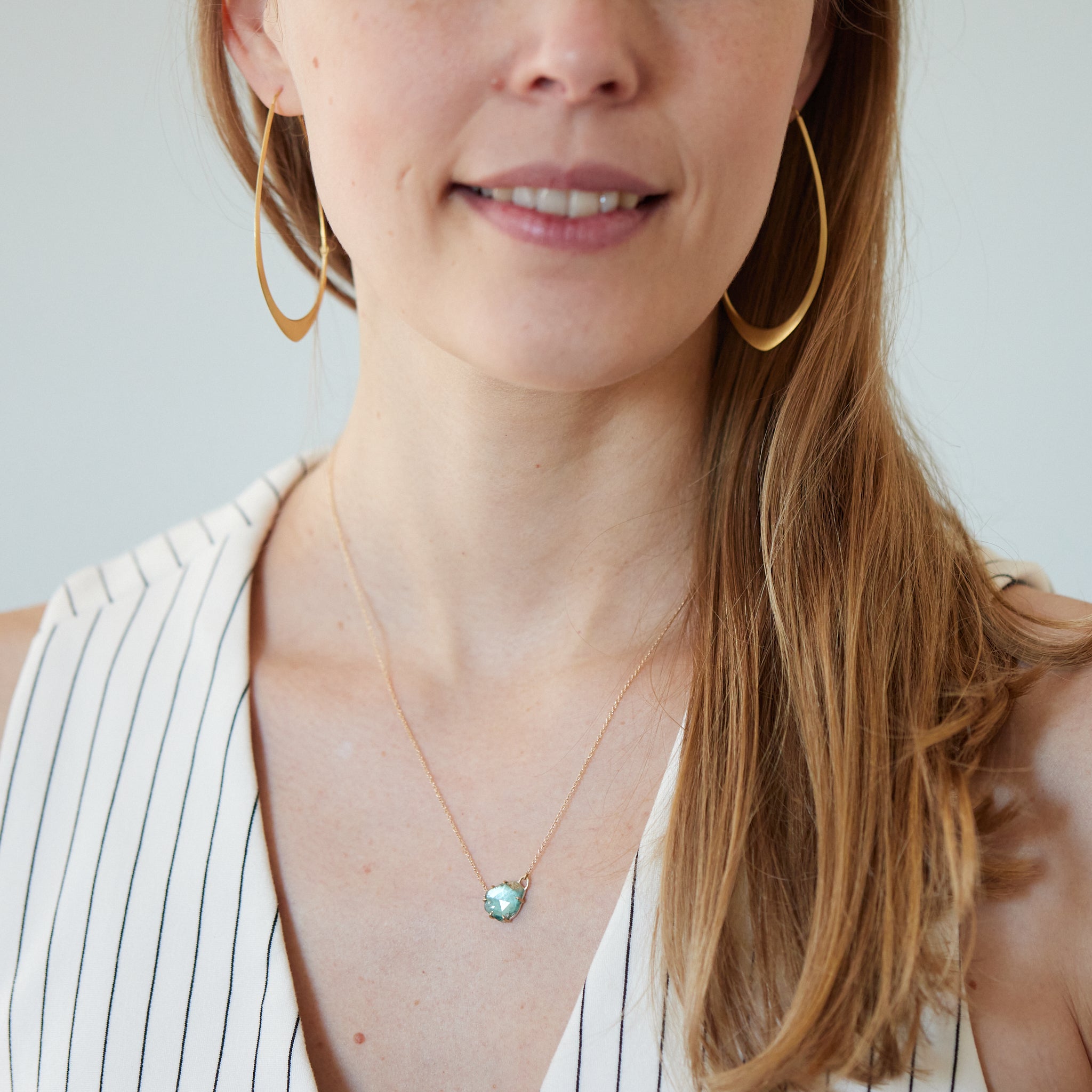 partially polished, partially raw blue-green tourmaline gemstone is set in gold prongs and has a gold chain attached to each side. Photographed on person.