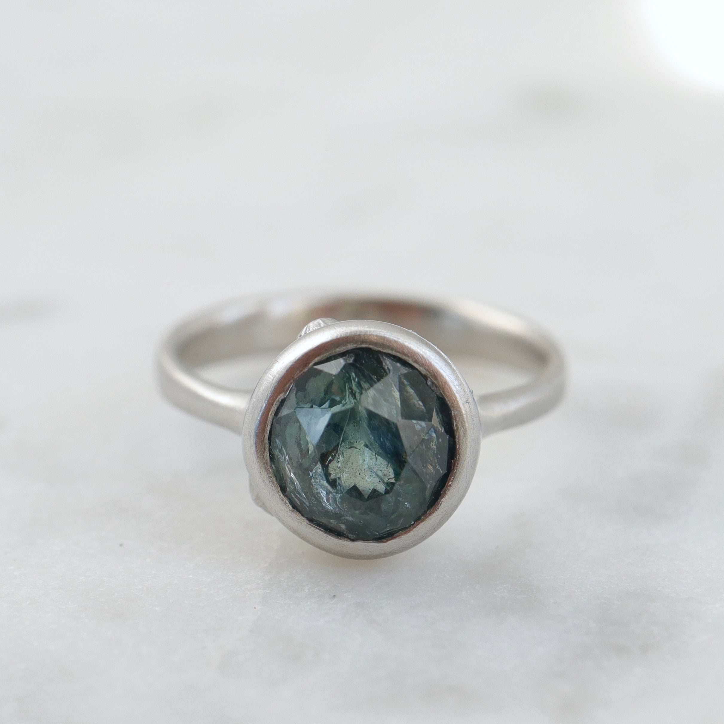 Front view of an included teal montana sapphire set in a platinum bezel and band. The ring has three tiny barnacles on the setting with diamonds within.