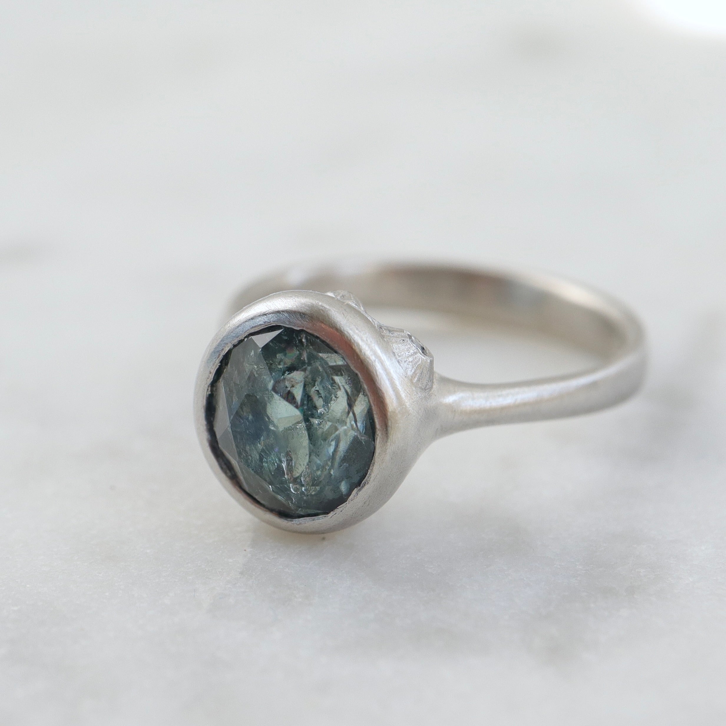 1/4 view of an included teal montana sapphire set in a platinum bezel and band. The ring has three tiny barnacles on the setting with diamonds within.