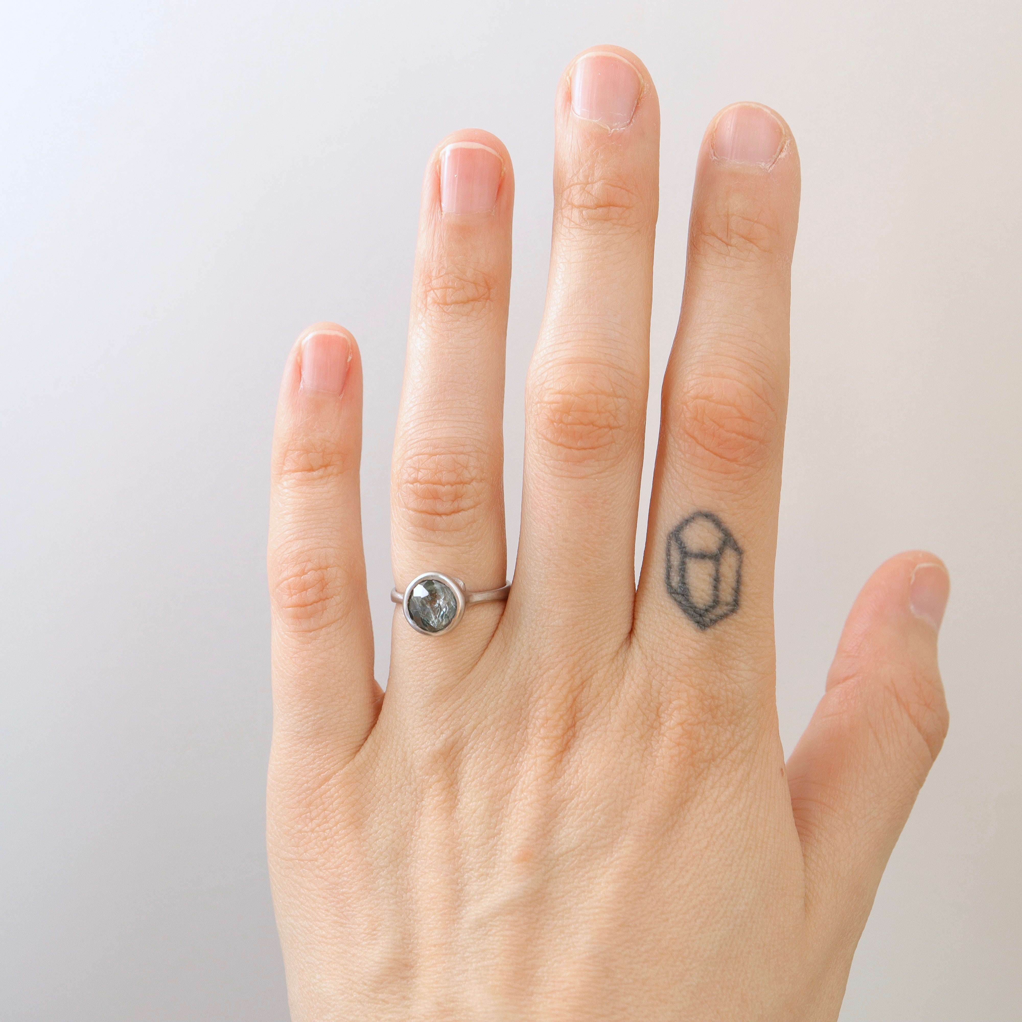On-hand view of an included teal montana sapphire set in a platinum bezel and band. The ring has three tiny barnacles on the setting with diamonds within.