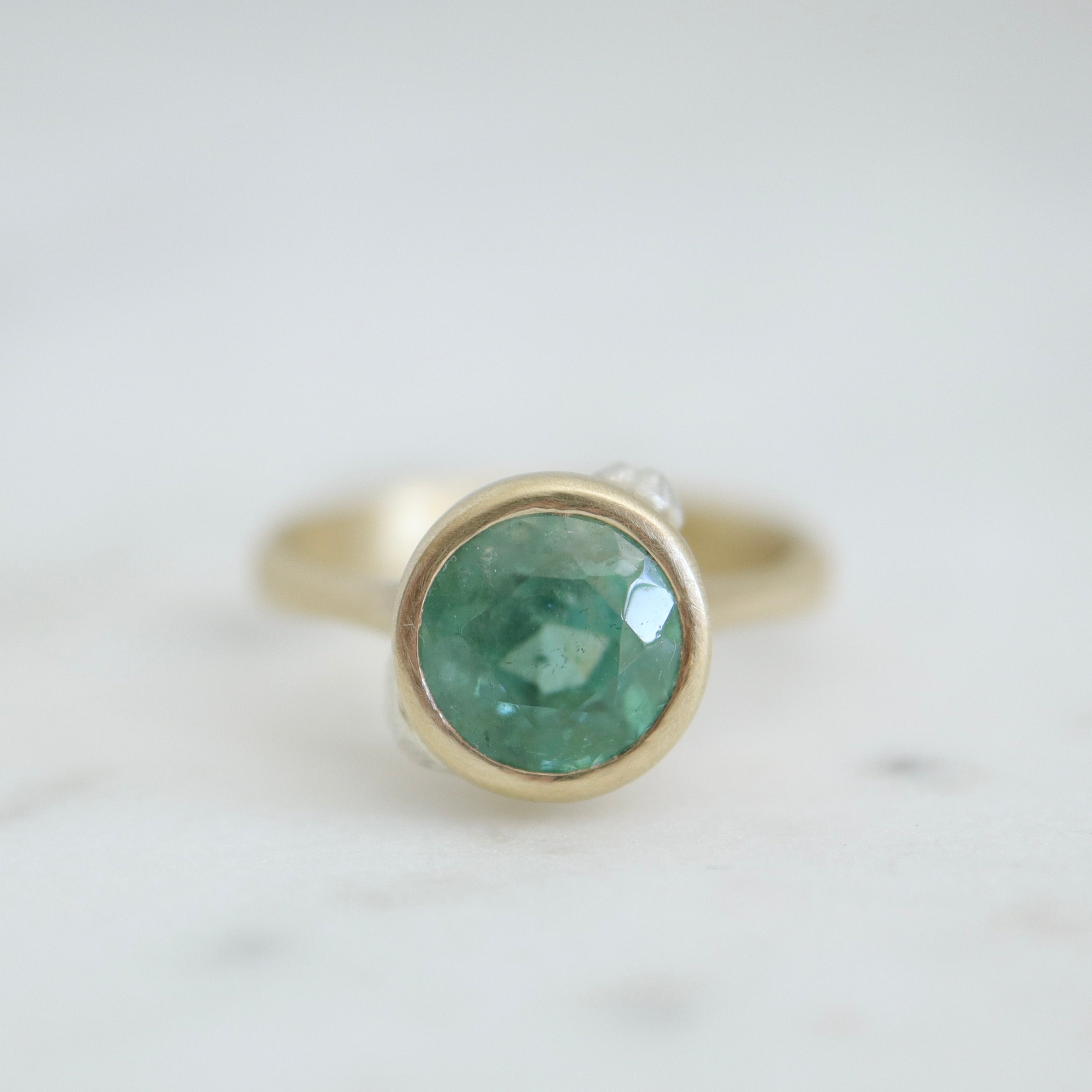 cyan tourmaline round stone set in a gold bezel setting. three little barnacles are soldered around the sides. Front view.
