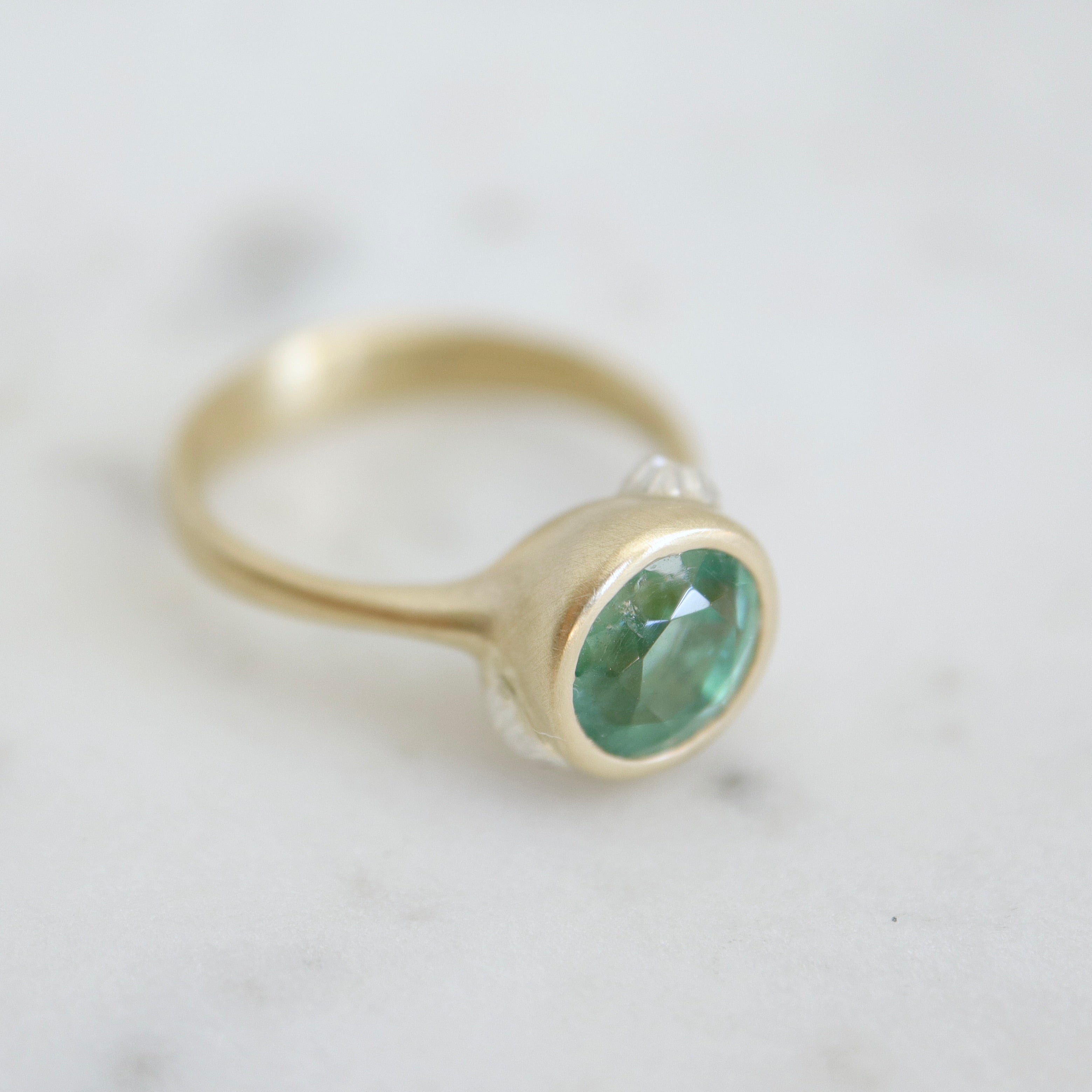 cyan tourmaline round stone set in a gold bezel setting. three little barnacles are soldered around the sides. 1/4 view.