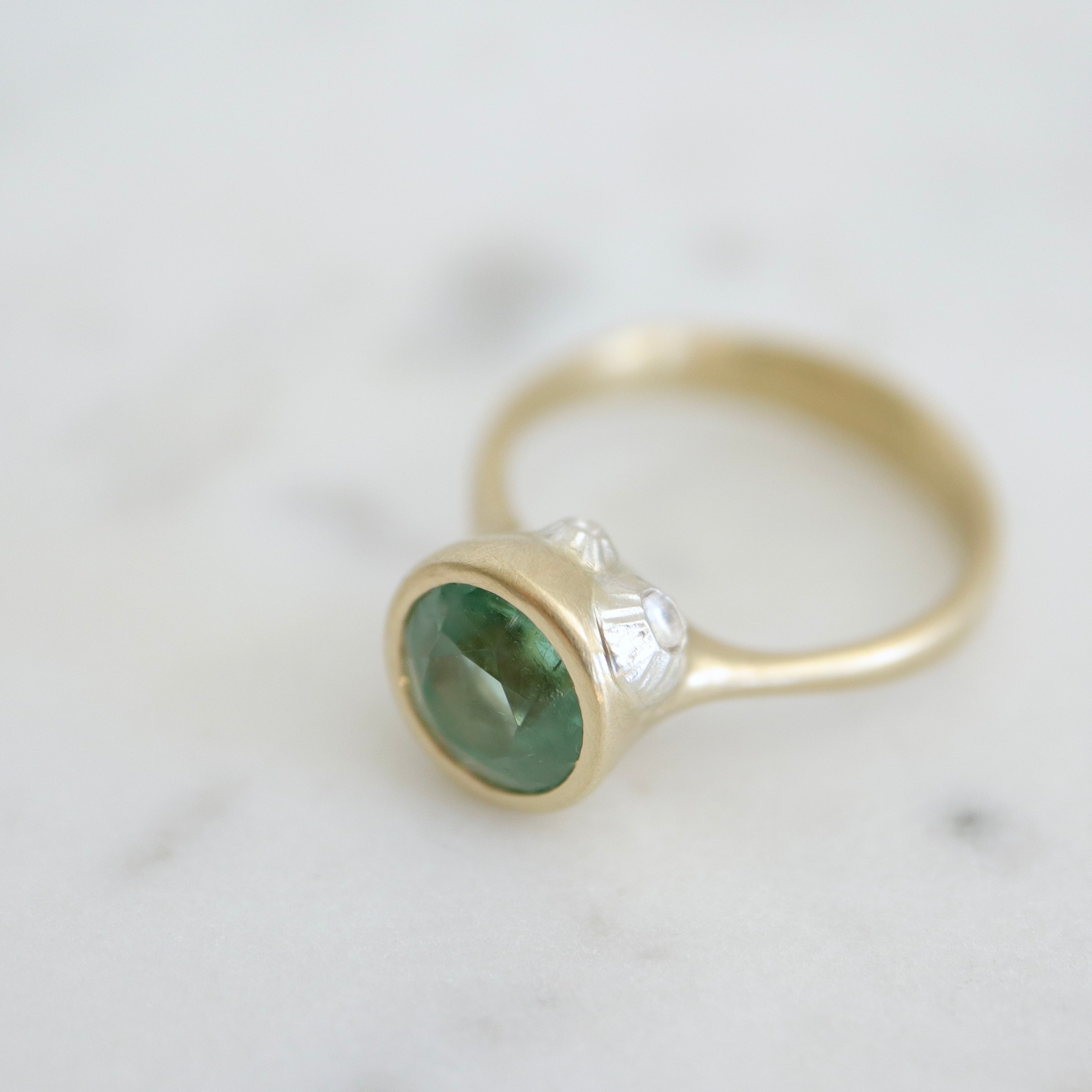 cyan tourmaline round stone set in a gold bezel setting. three little barnacles are soldered around the sides. 1/4 view.