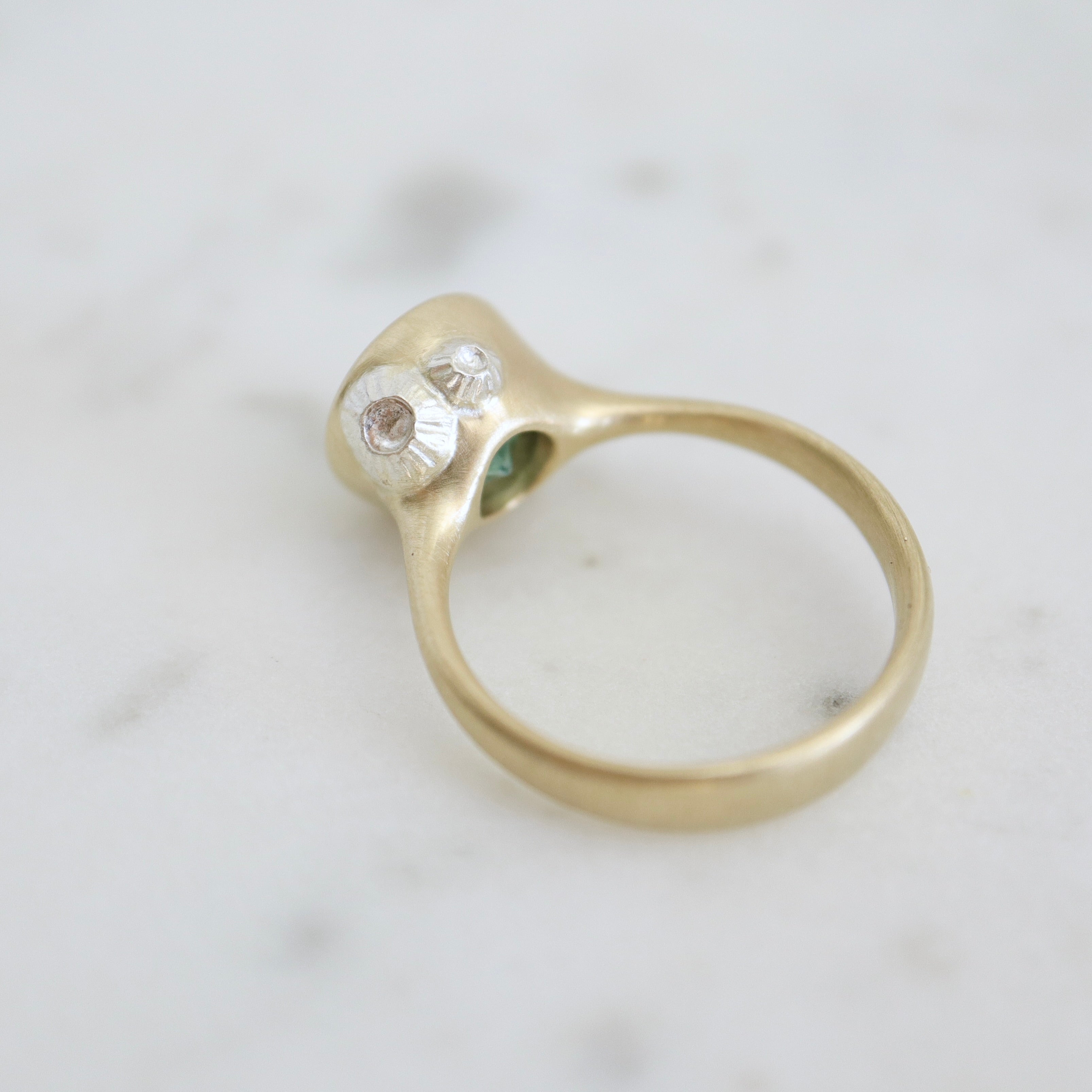 cyan tourmaline round stone set in a gold bezel setting. three little barnacles are soldered around the sides. Back view.