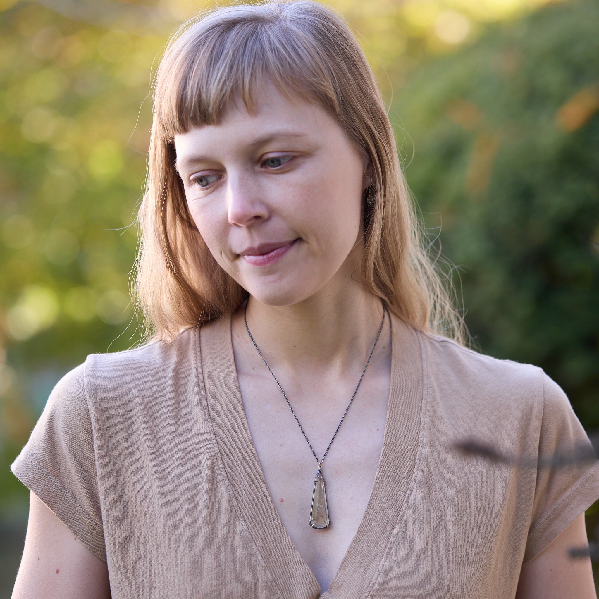 Woman wearing a beige teeshirt with a long pendant necklace with a blurred natural background