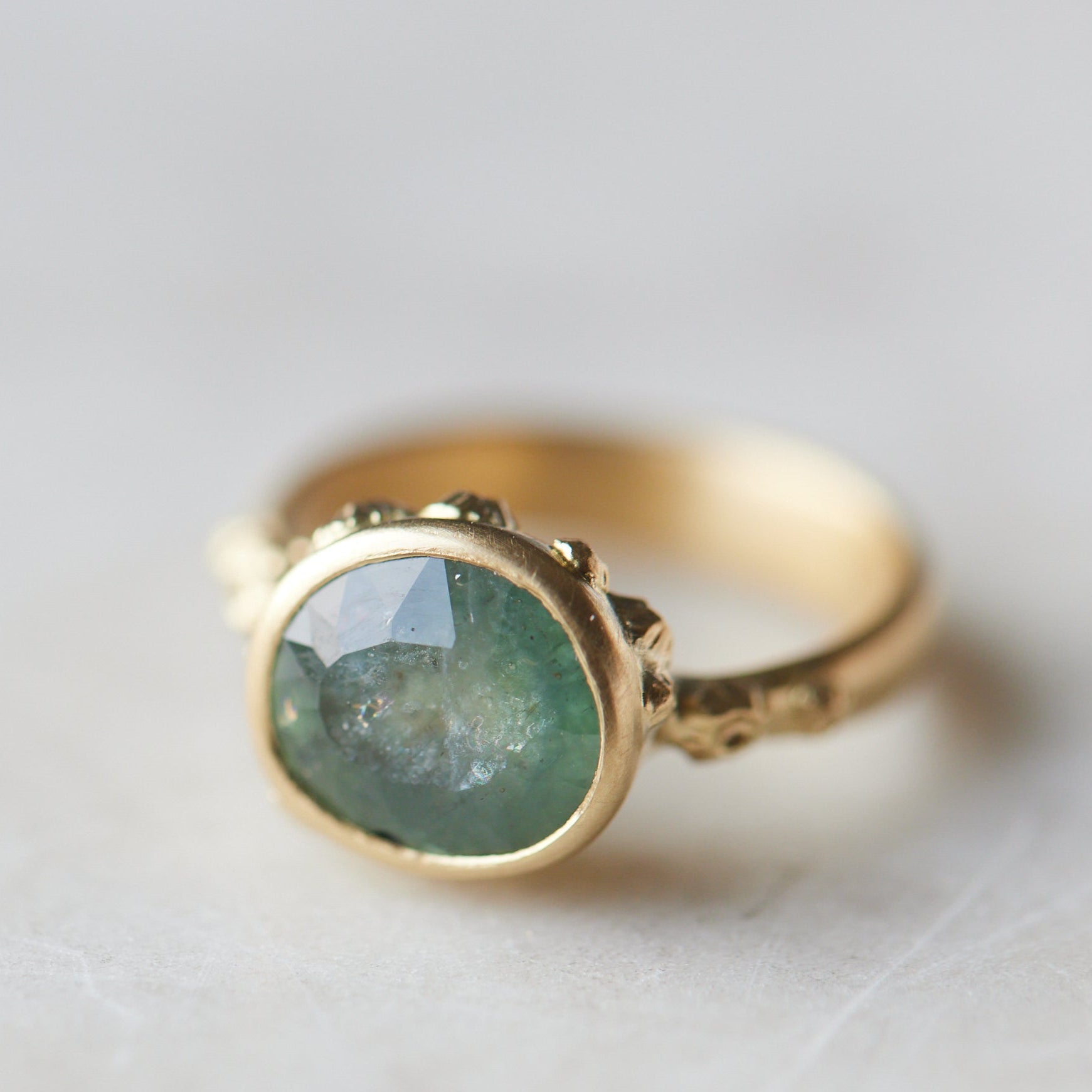 aqua colored teal montana sapphire stone set in a bezel setting of a gold ring with barnacles in a nautical aesthetic. Photographed at 1/4 view, with a marble background.