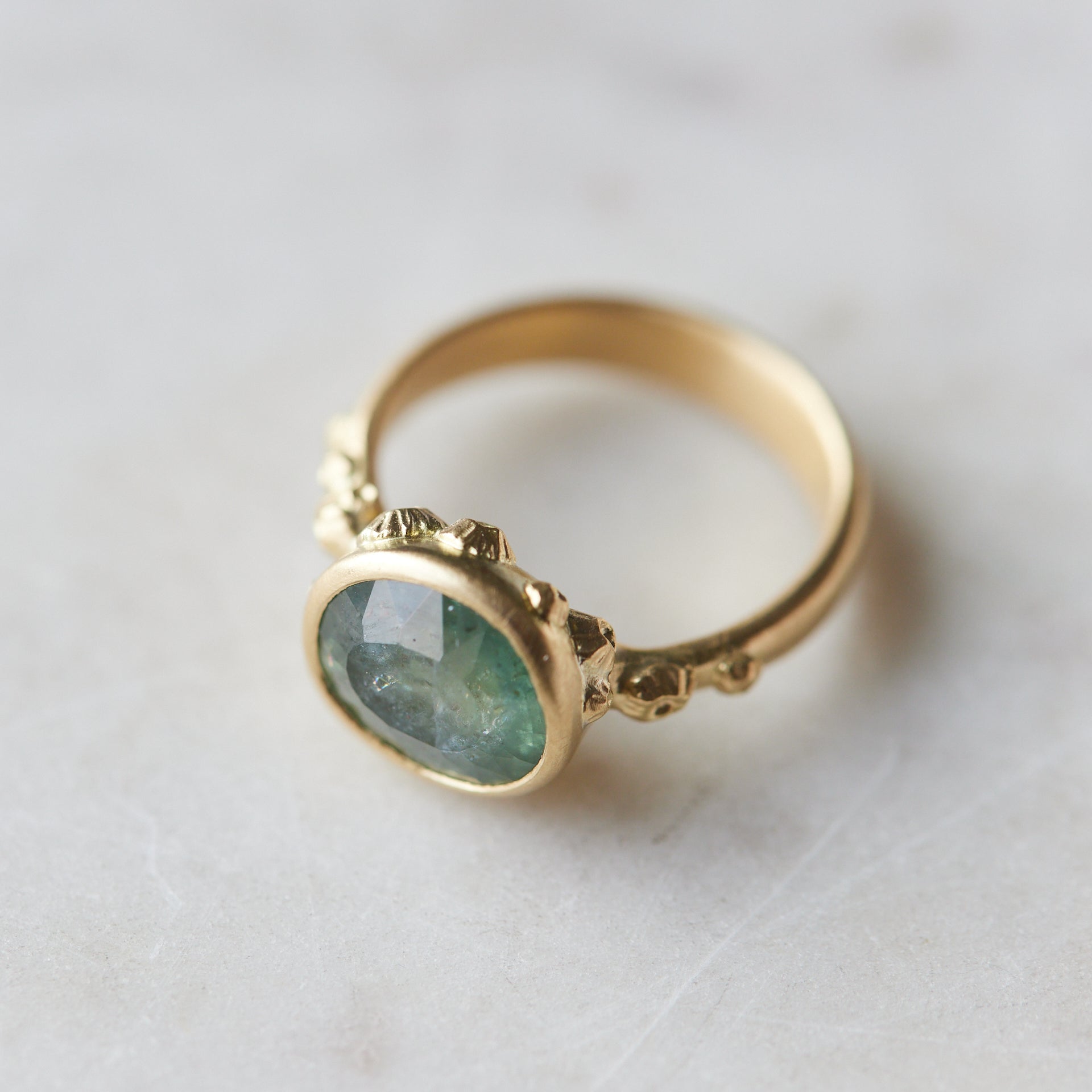 aqua colored teal montana sapphire stone set in a bezel setting of a gold ring with barnacles in a nautical aesthetic. Photographed at 1/4 view, with a marble background.