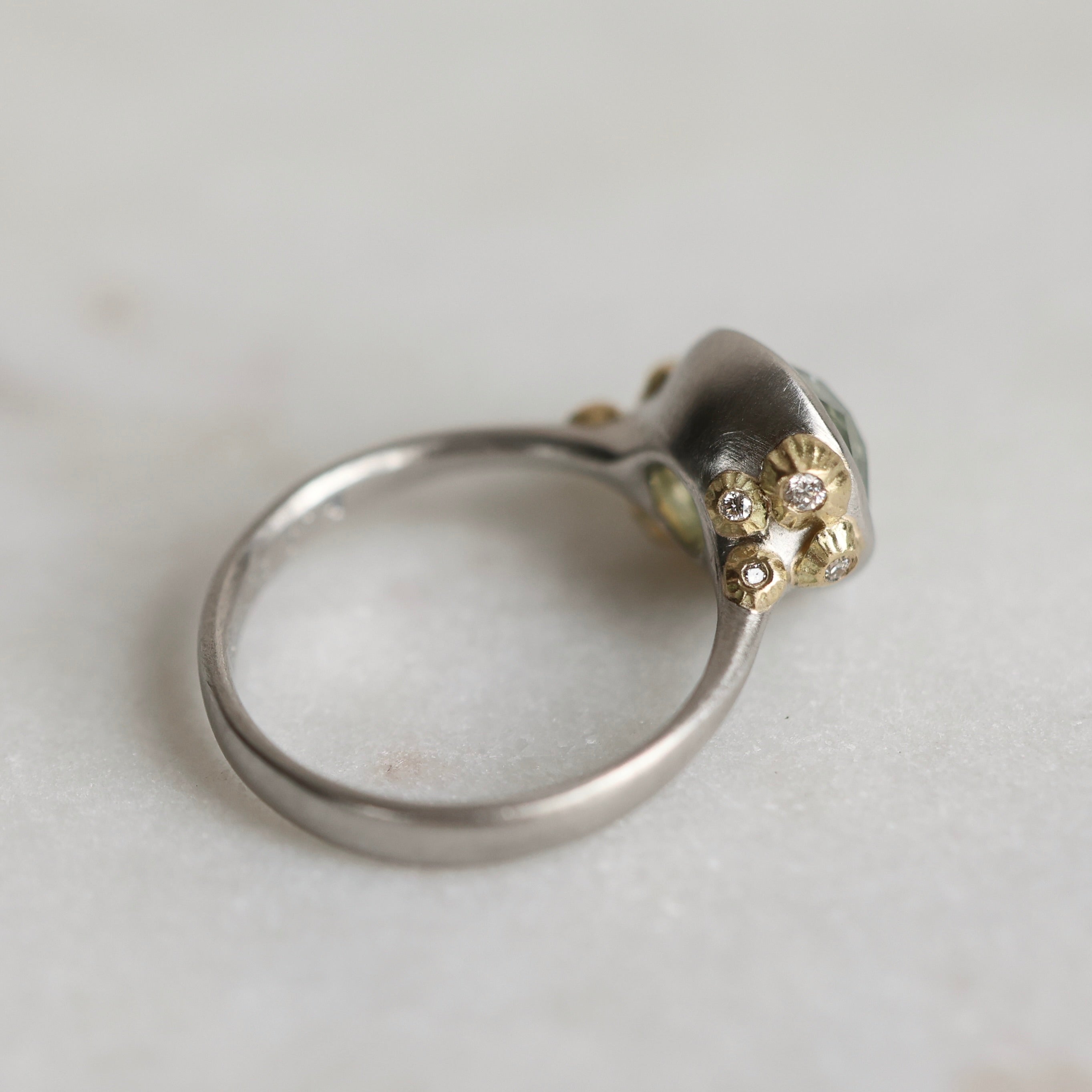 Ring with a green gemstone and platinum band with yellow gold  and diamond details on a light gray background