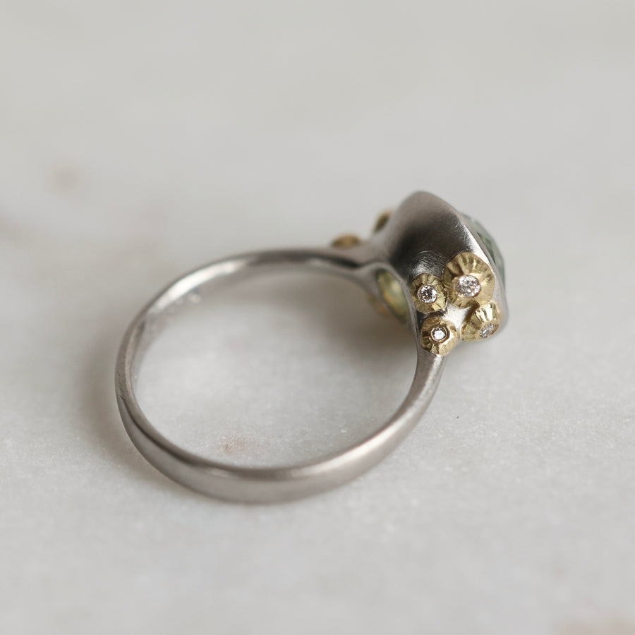 Ring with a green gemstone and platinum band with yellow gold  and diamond details on a light gray background
