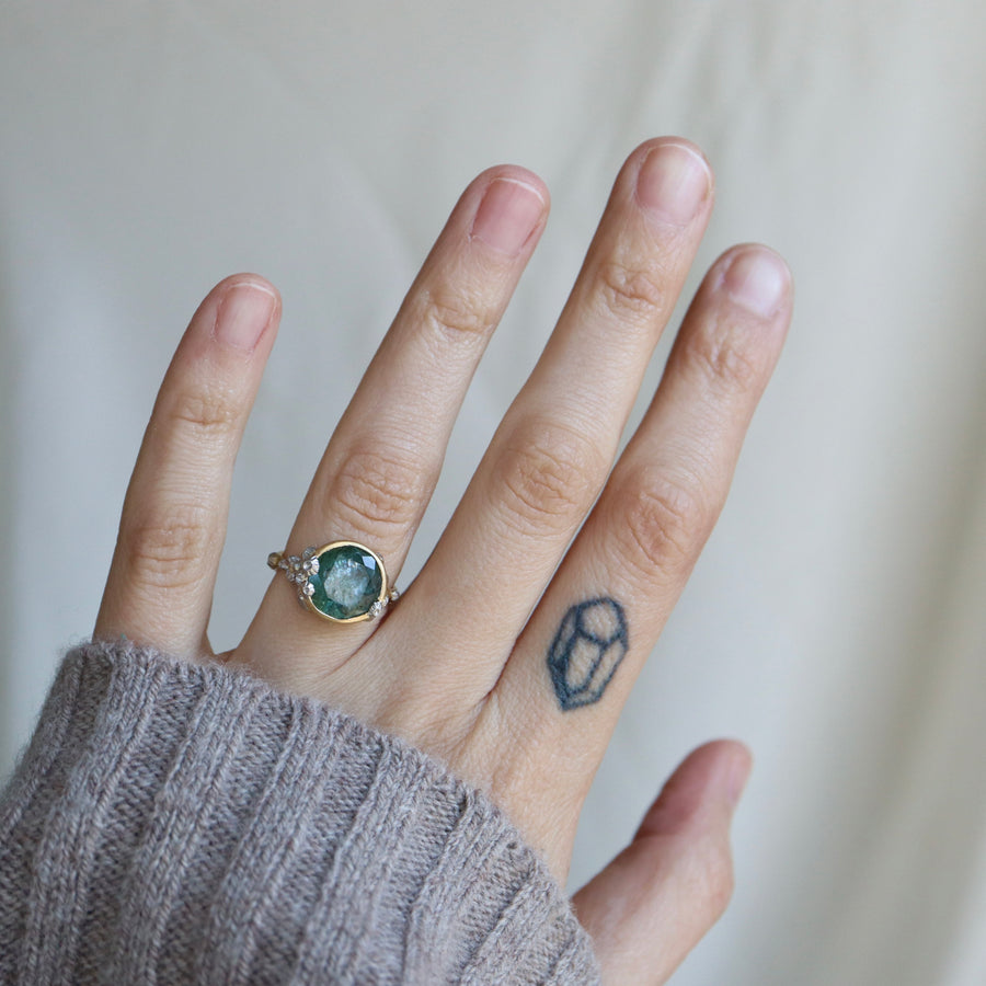 hand wearing a teal gemstone and gold ring with barnacles against a pale background