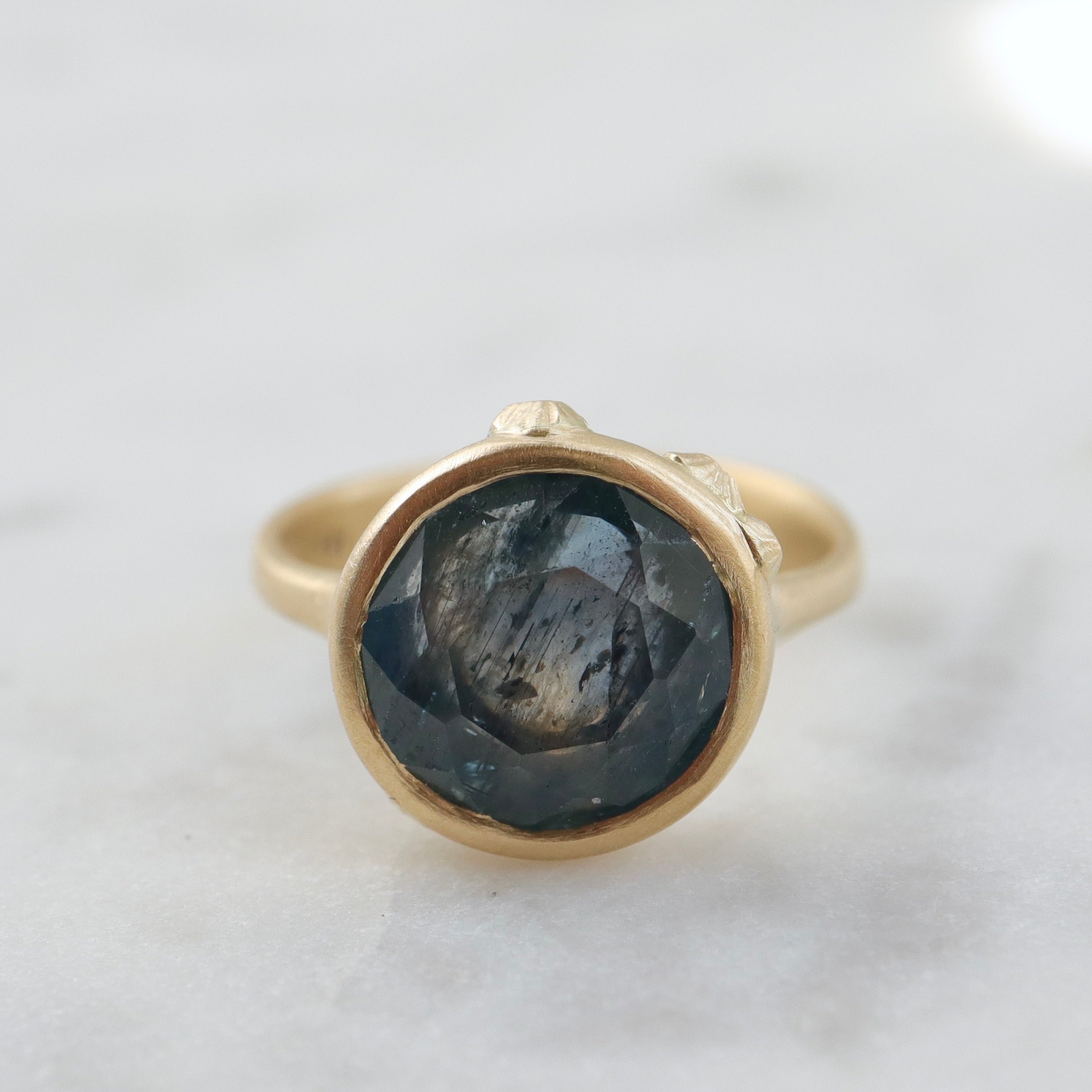 steely blue color change sapphire set in a gold bezel and band. Around the cup of the bezel are tiny gold barnacles. Photographed front view and in natural light.