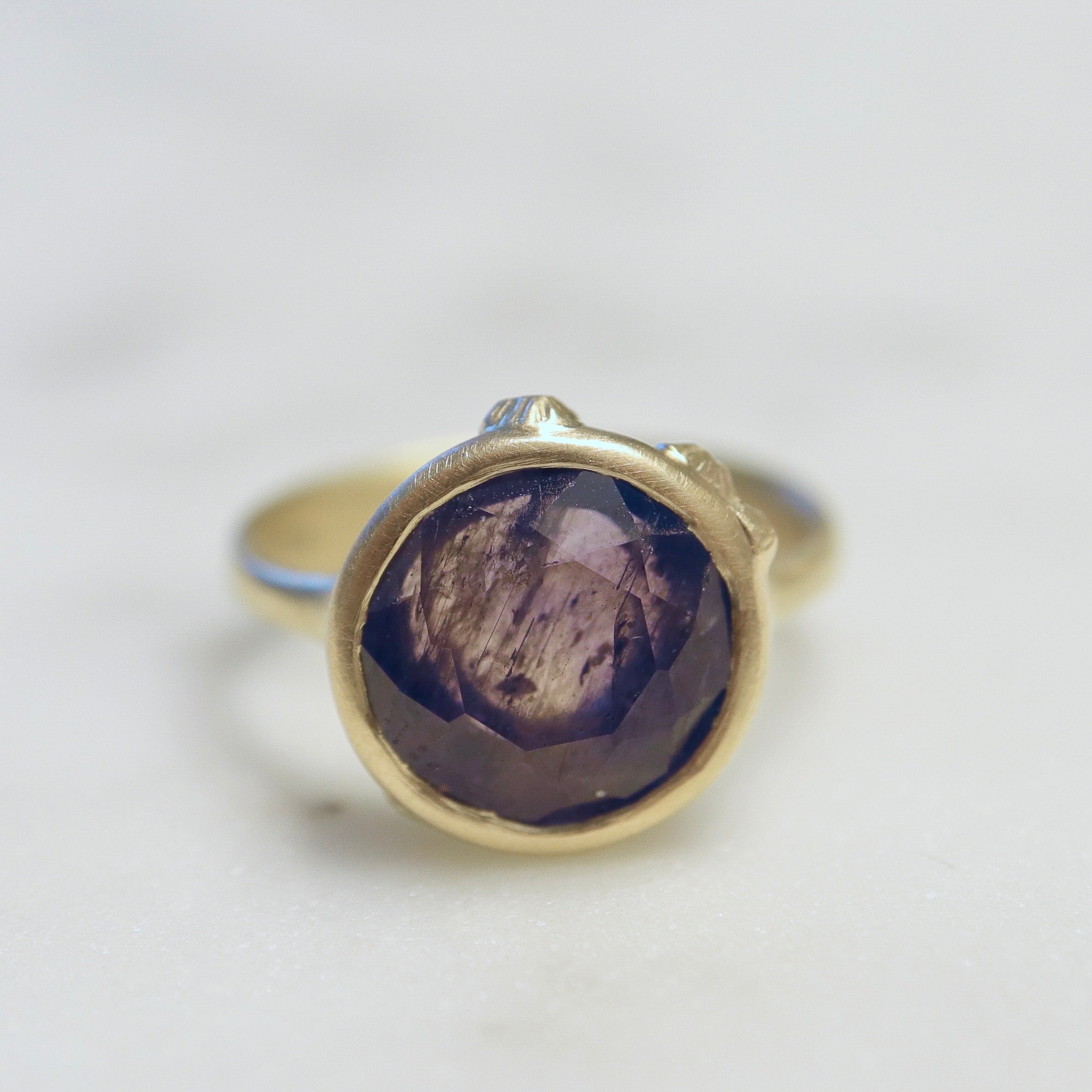 mulberry color change sapphire set in a gold bezel and band. Around the cup of the bezel are tiny gold barnacles. Photographed front view and in artificial light.