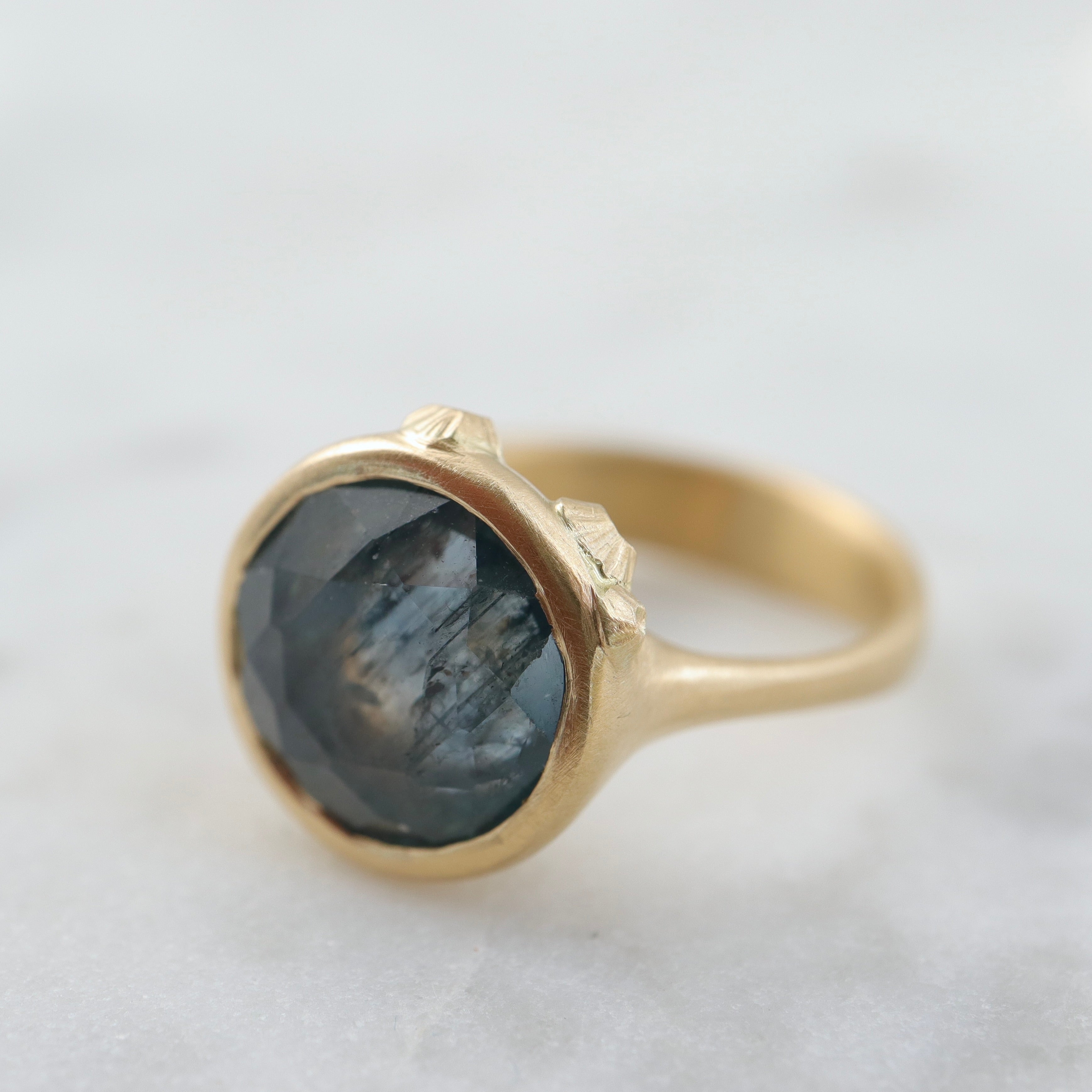 steely blue color change sapphire set in a gold bezel and band. Around the cup of the bezel are tiny gold barnacles. Photographed side view and in natural light.