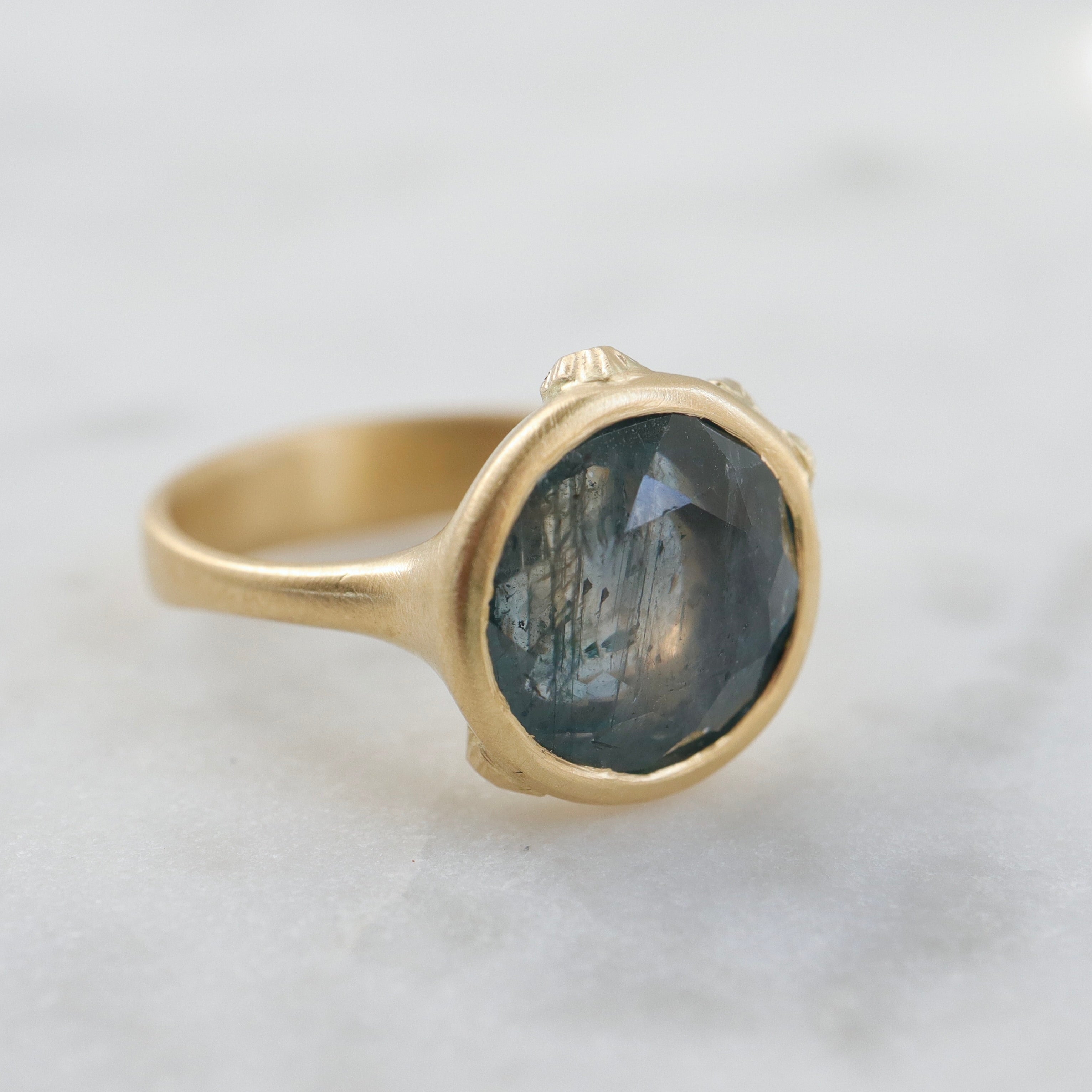 steely blue color change sapphire set in a gold bezel and band. Around the cup of the bezel are tiny gold barnacles. Photographed side view and in natural light.