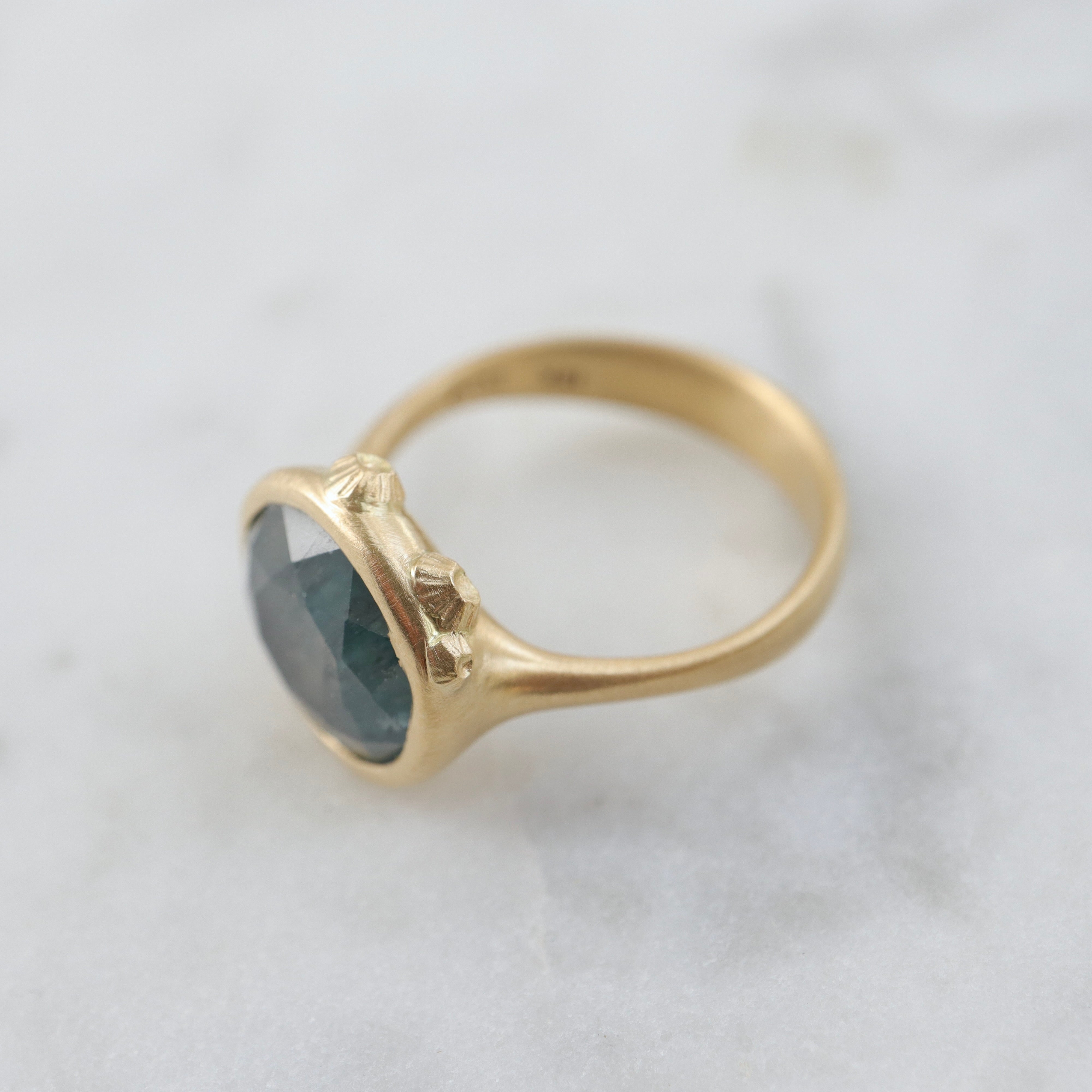 steely blue color change sapphire set in a gold bezel and band. Around the cup of the bezel are tiny gold barnacles. Photographed front side and in natural light.