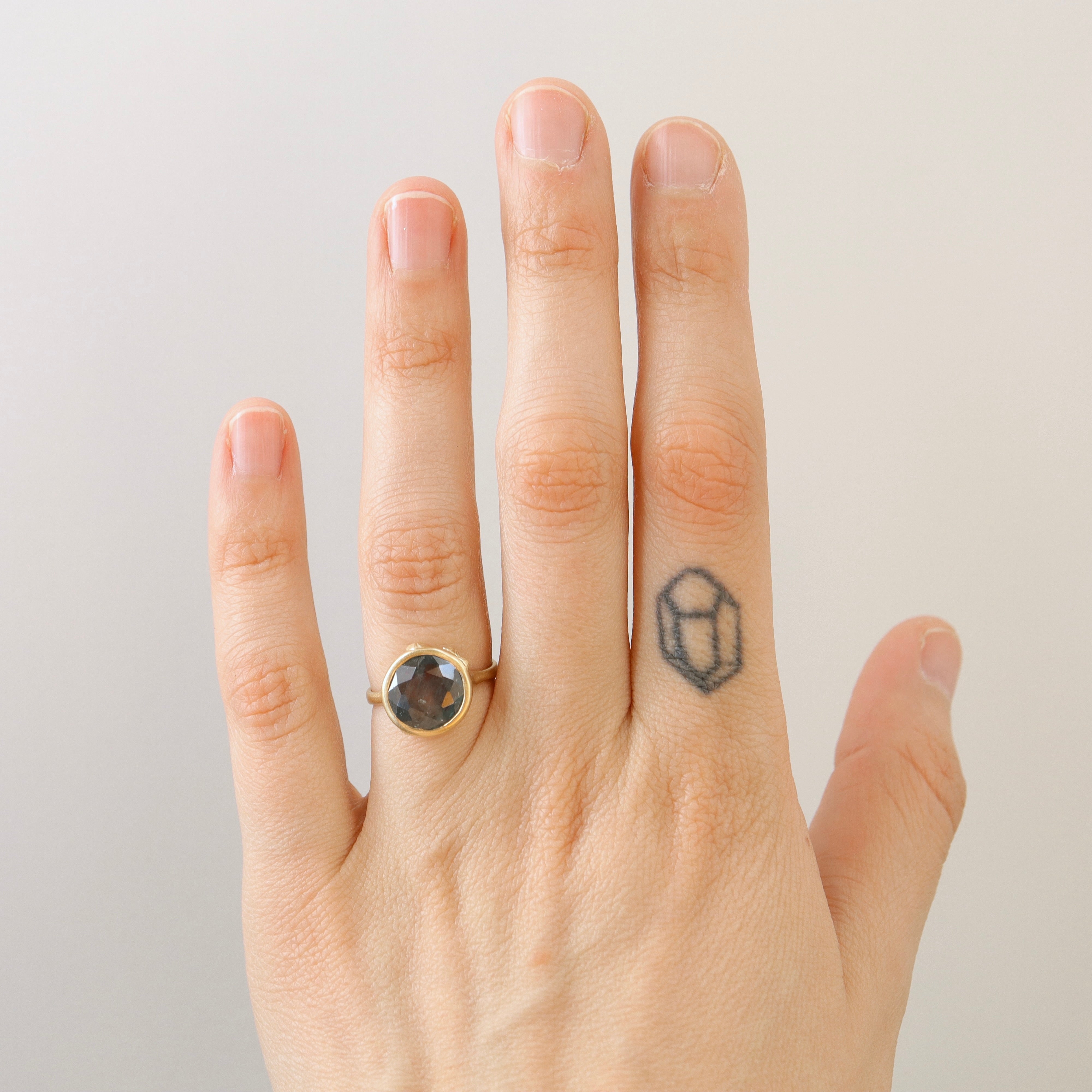 steely blue color change sapphire set in a gold bezel and band. Around the cup of the bezel are tiny gold barnacles. Photographed on hand and in natural light.