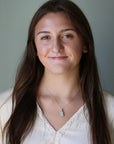 Woman with long brown hair wearing a white blouse and a necklace against a plain background