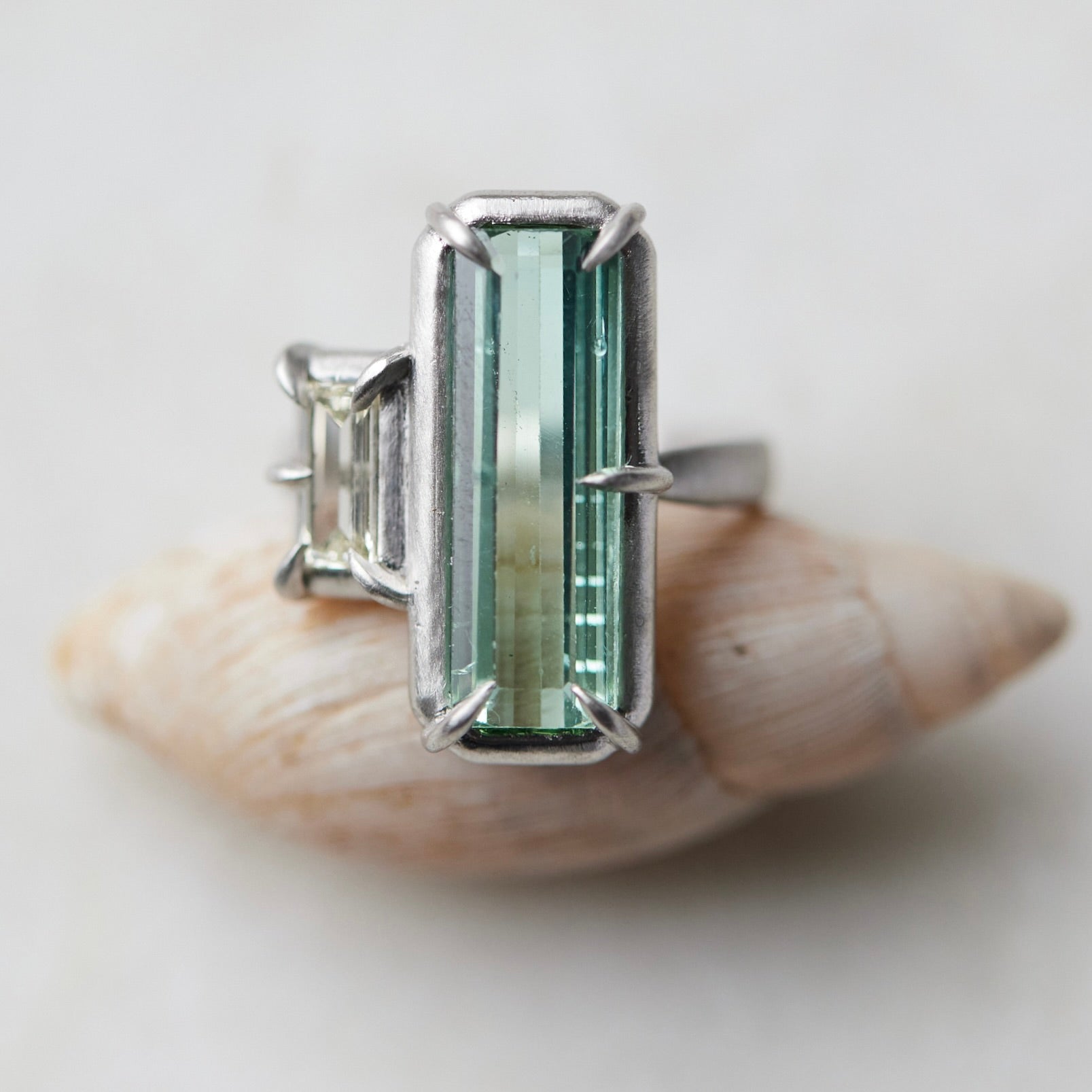 long, rectangular tourmaline stone set with prongs next to a diamond baguette. The ring is platinum, and shown balancing on a shell.