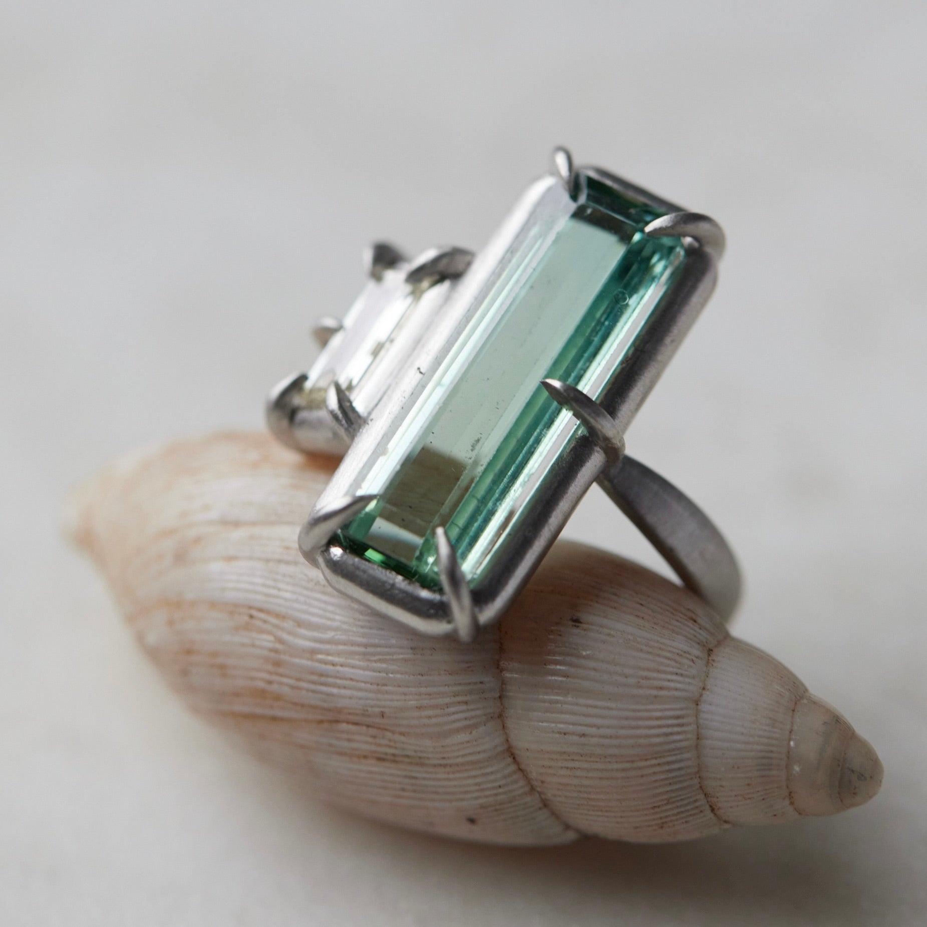 long, rectangular tourmaline stone set with prongs next to a diamond baguette. The ring is platinum, and shown balancing on a shell.