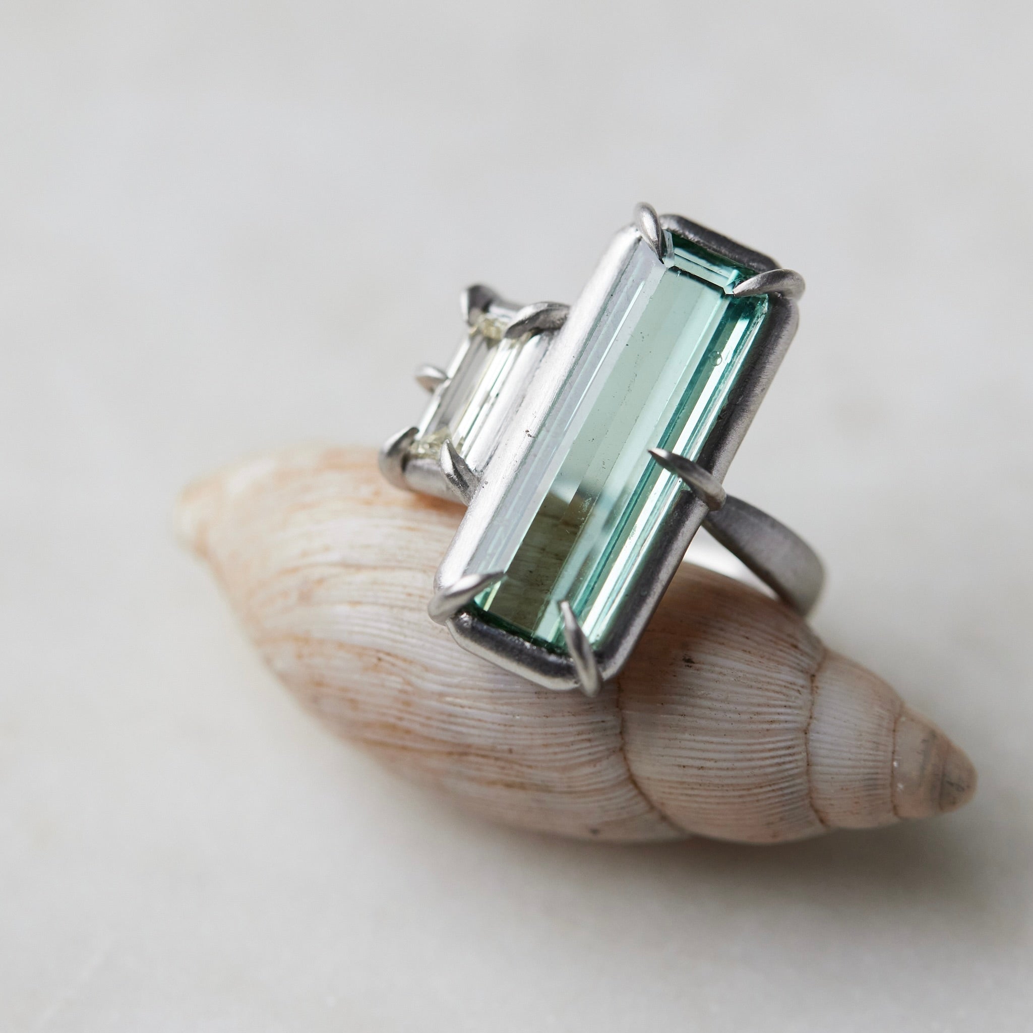 long, rectangular tourmaline stone set with prongs next to a diamond baguette. The ring is platinum, and shown balancing on a shell.