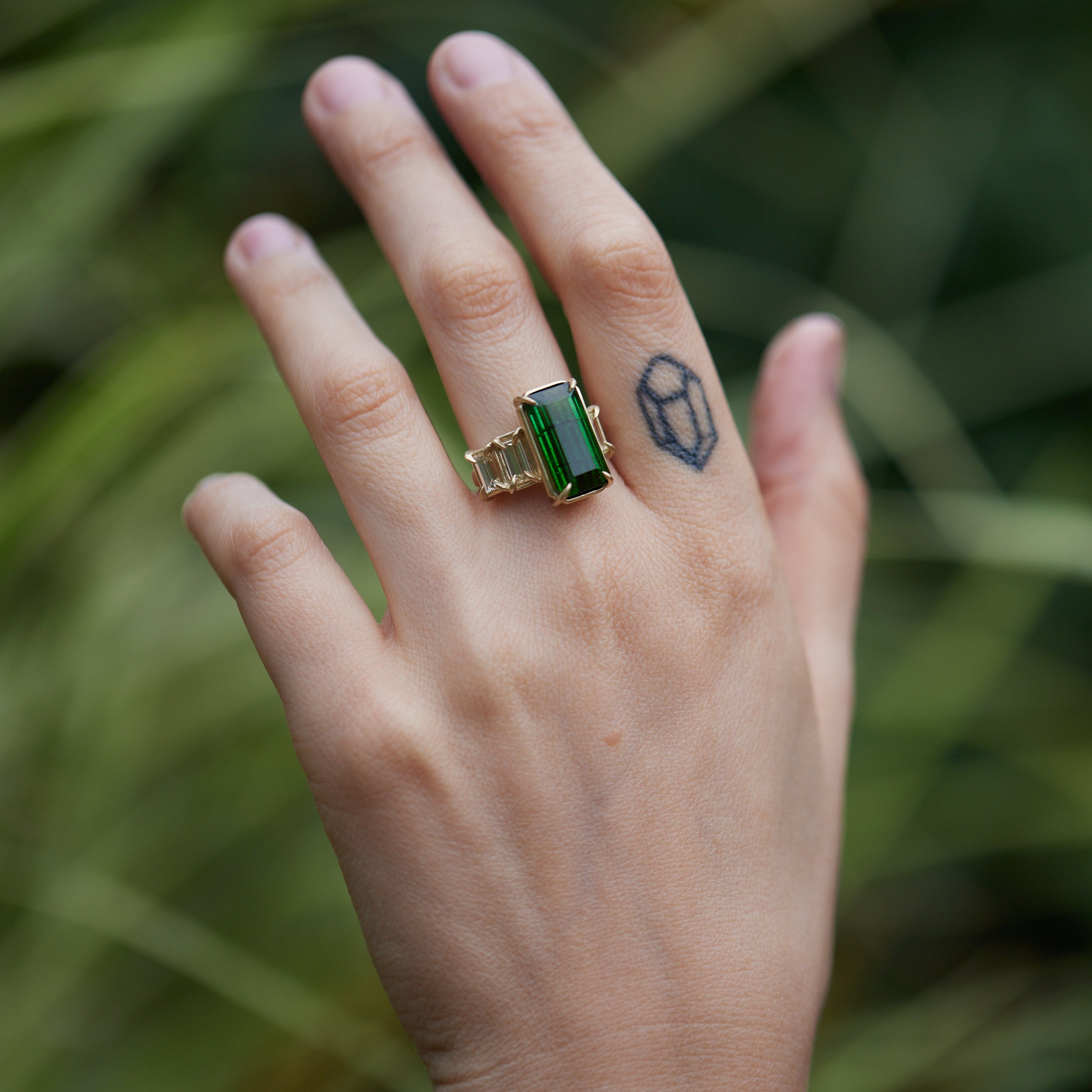 a grand center rectangular green tourmaline gemstone is set within four prongs. On one side is one diamond baguette, on the other sit two. The four stones make an off-set centerpiece of a gold ring. Shown on hand.