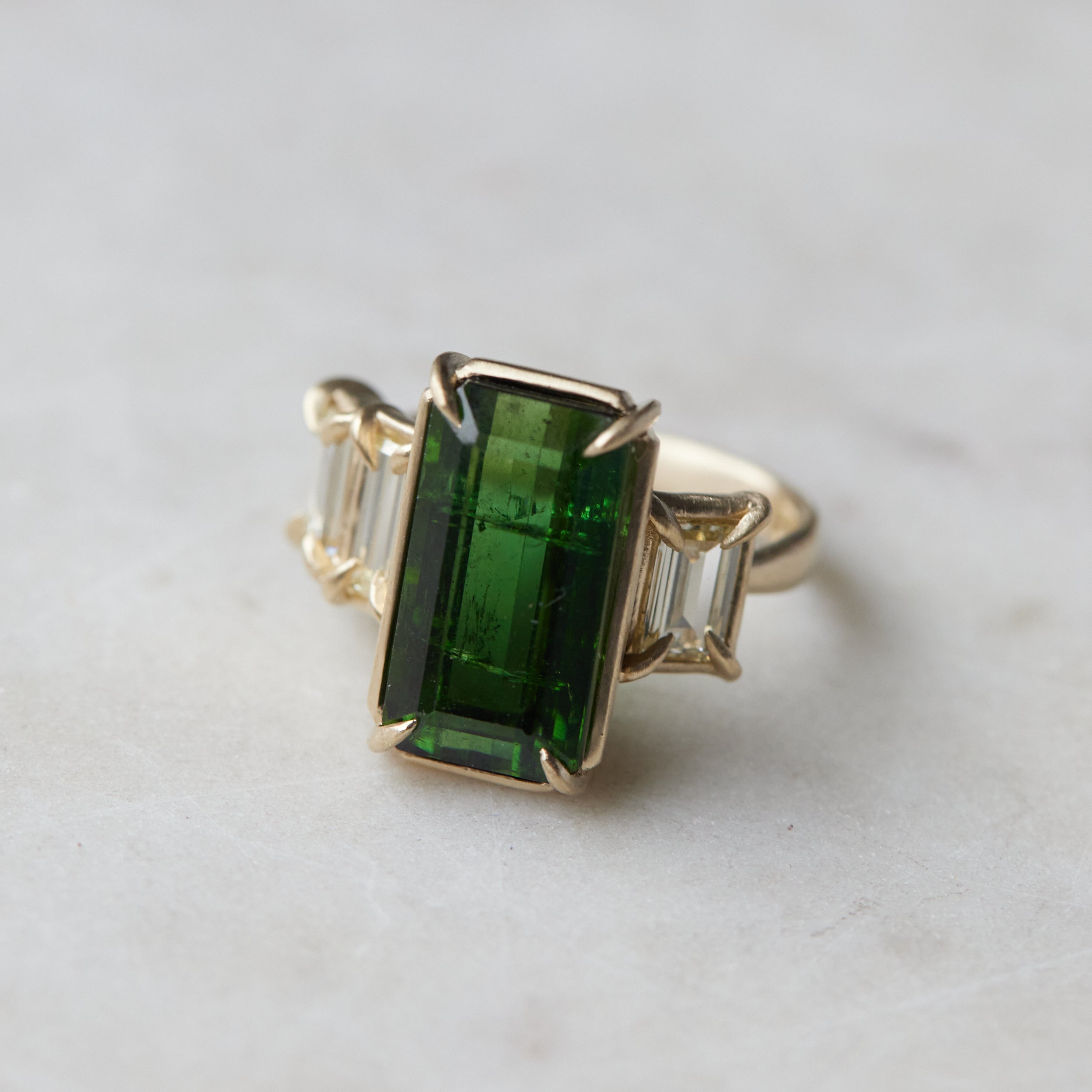 a grand center rectangular green tourmaline gemstone is set within four prongs. On one side is one diamond baguette, on the other sit two. The four stones make an off-set centerpiece of a gold ring. 