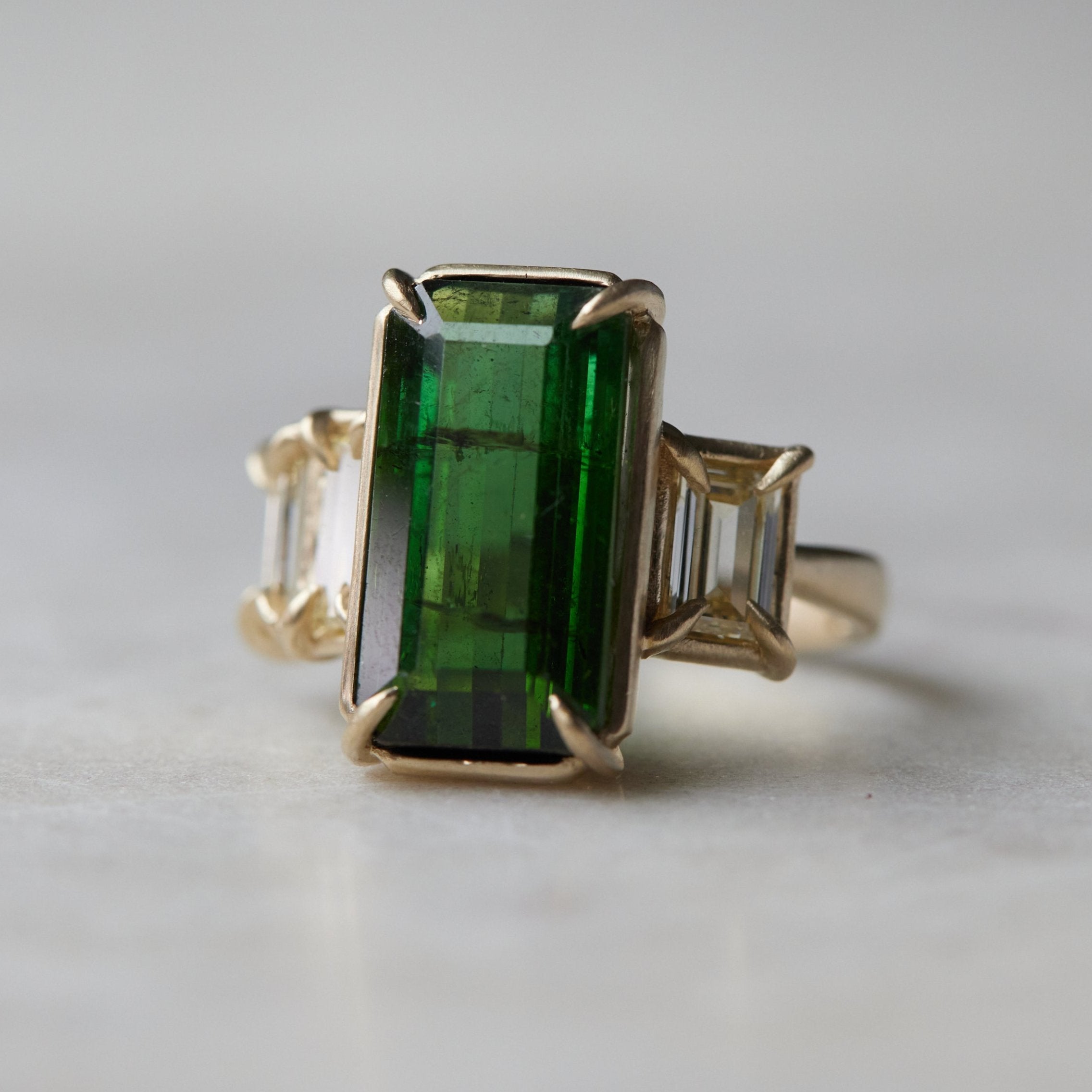 a grand center rectangular green tourmaline gemstone is set within four prongs. On one side is one diamond baguette, on the other sit two. The four stones make an off-set centerpiece of a gold ring.