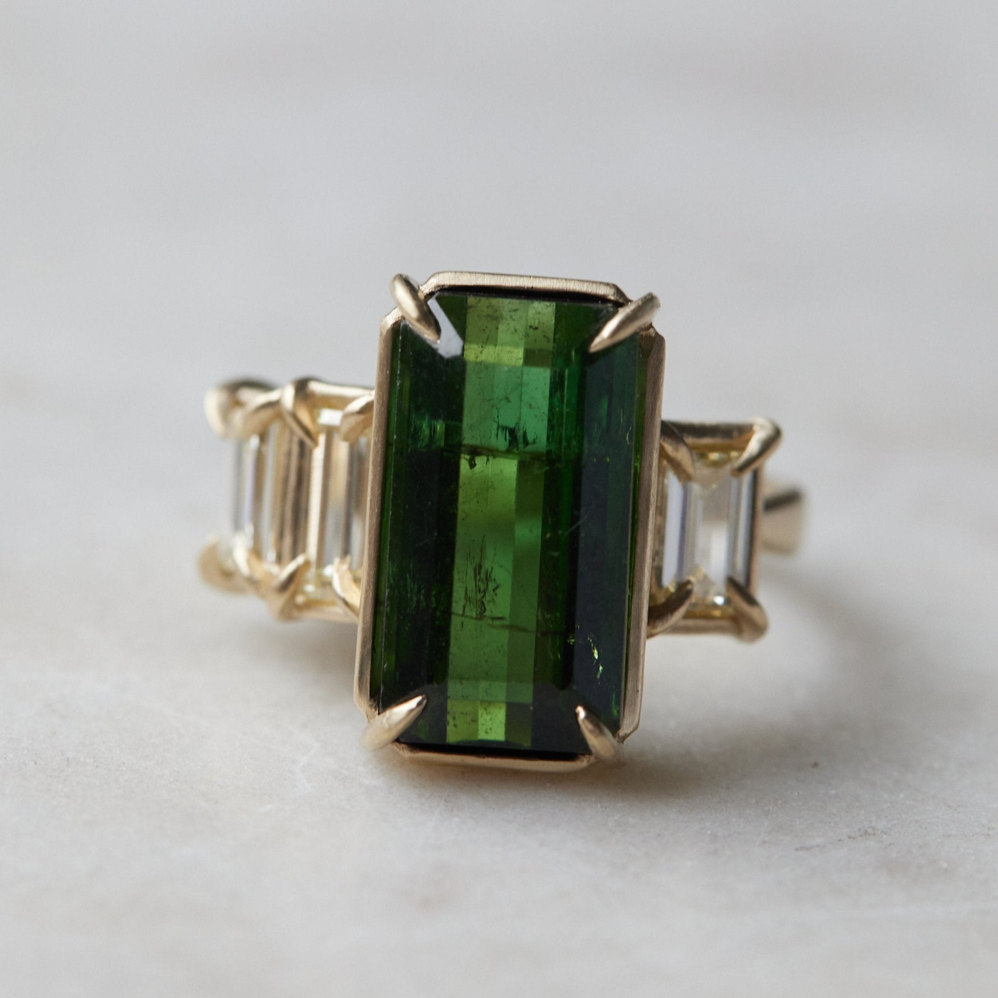 a grand center rectangular green tourmaline gemstone is set within four prongs. On one side is one diamond baguette, on the other sit two. The four stones make an off-set centerpiece of a gold ring.