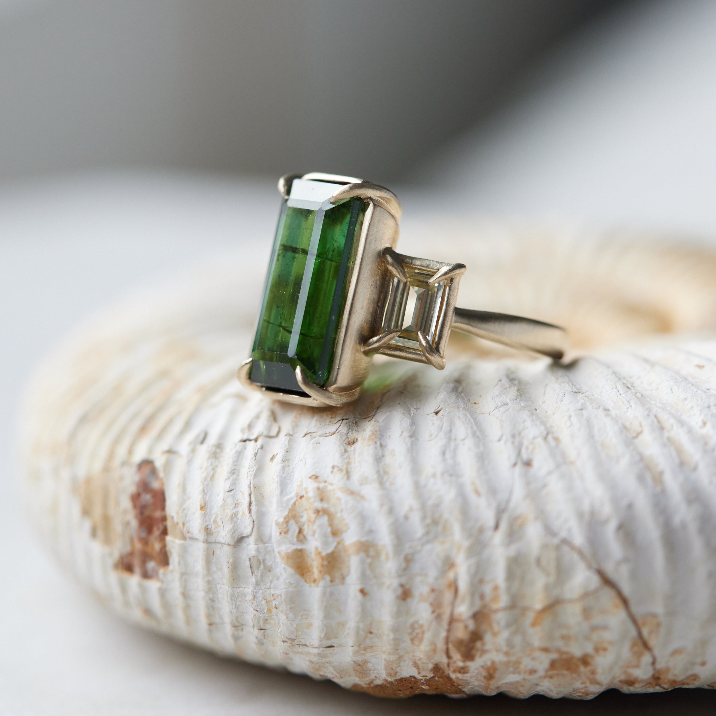 a grand center rectangular green tourmaline gemstone is set within four prongs. On one side is one diamond baguette, on the other sit two. The four stones make an off-set centerpiece of a gold ring. Shown on an ammonite fossil.