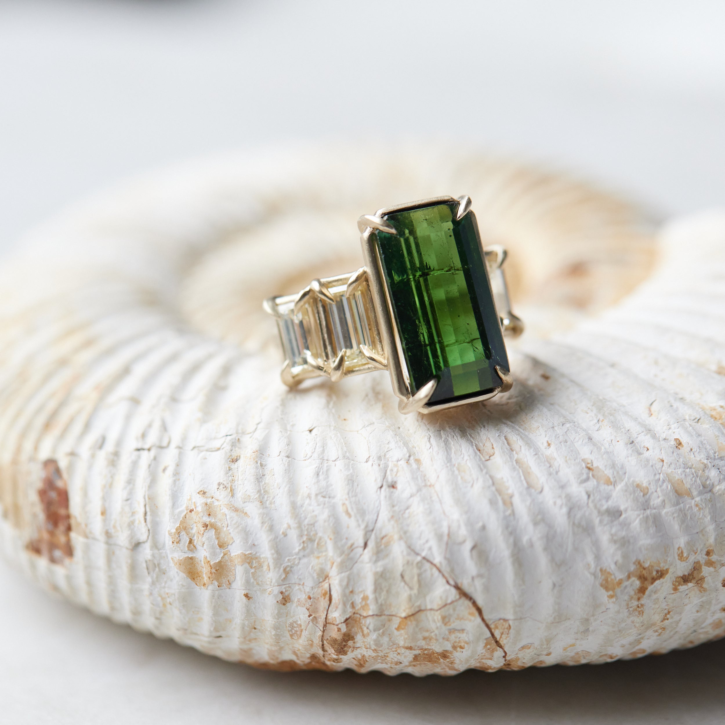 a grand center rectangular green tourmaline gemstone is set within four prongs. On one side is one diamond baguette, on the other sit two. The four stones make an off-set centerpiece of a gold ring. Shown on an ammonite fossil.