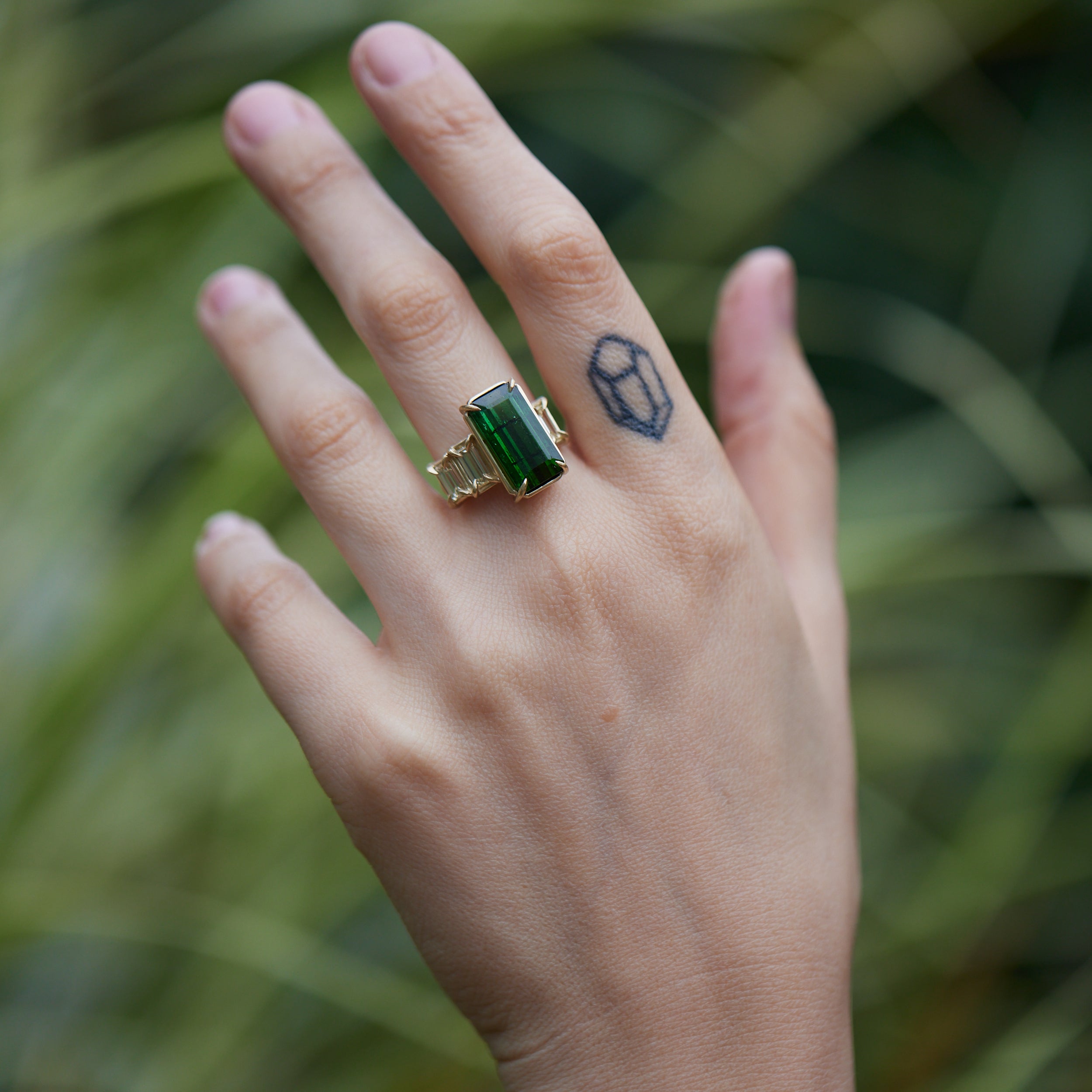 a grand center rectangular green tourmaline gemstone is set within four prongs. On one side is one diamond baguette, on the other sit two. The four stones make an off-set centerpiece of a gold ring. Shown on hand.