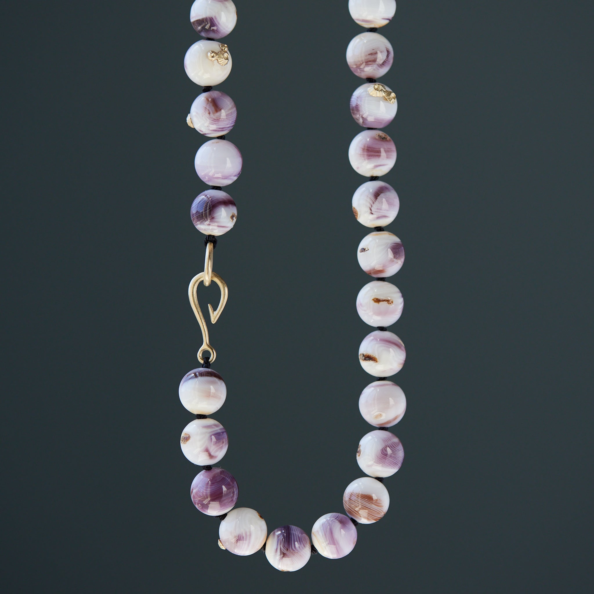 purple and white swirly beads of round quahog shells are strung on black silk and have gold barnacles attached throughout. A gold fish hook clasp attached the strand. Detail shot on dark grey background