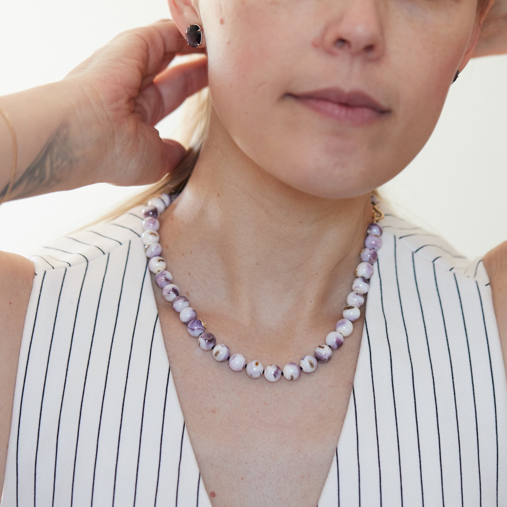 purple and white swirly beads of round quahog shells are strung on black silk and have gold barnacles attached throughout. A gold fish hook clasp attached the strand. Photographed on person.