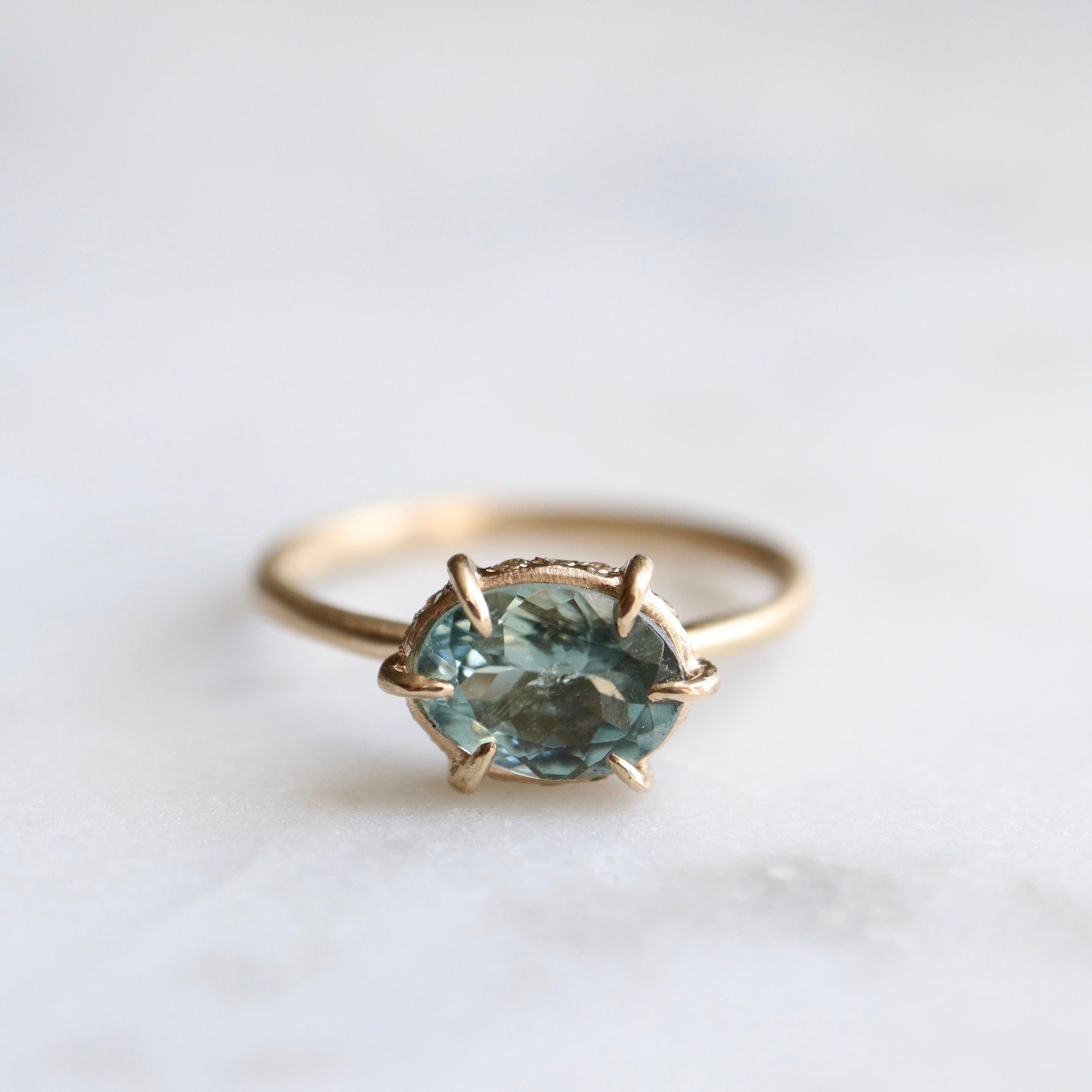 teal oval tourmaline stone set in a gold ring with gold prongs and diamonds set around the seat.