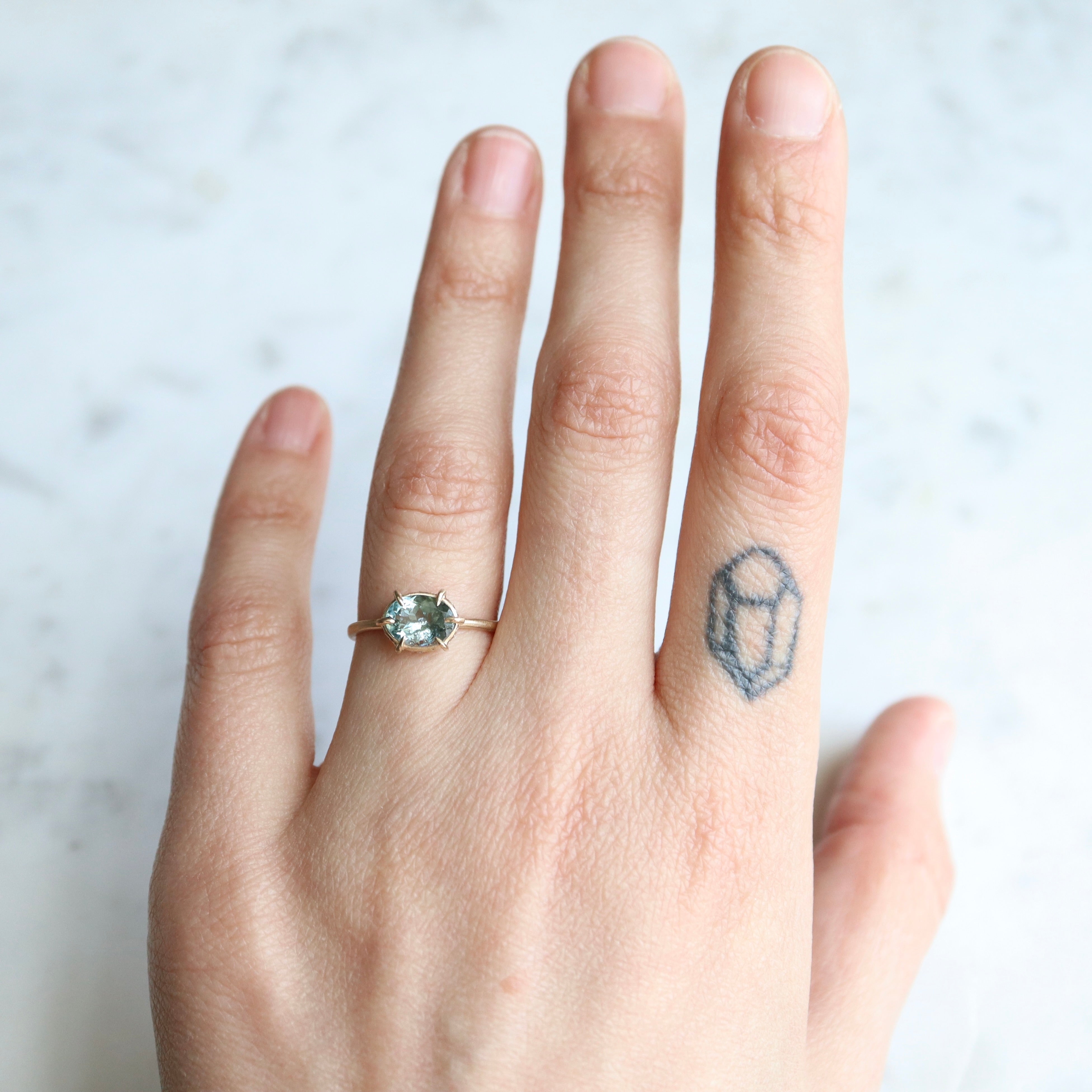 teal oval tourmaline stone set in a gold ring with gold prongs and diamonds set around the seat. on hand.