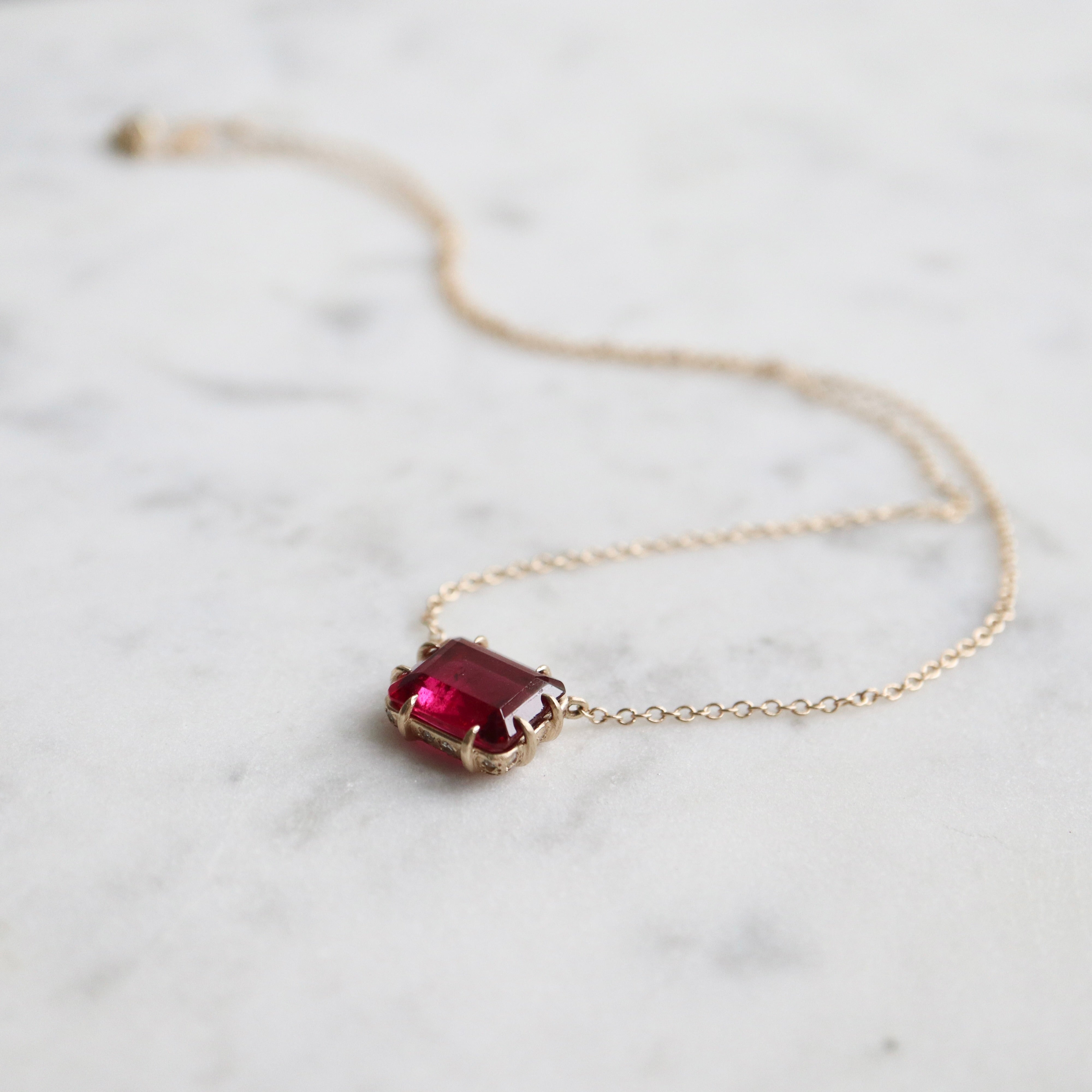 pink rectangular stone with in gold prongs on a gold chain with diamonds set in a hidden halo on the side of the setting. photographed on white marble.