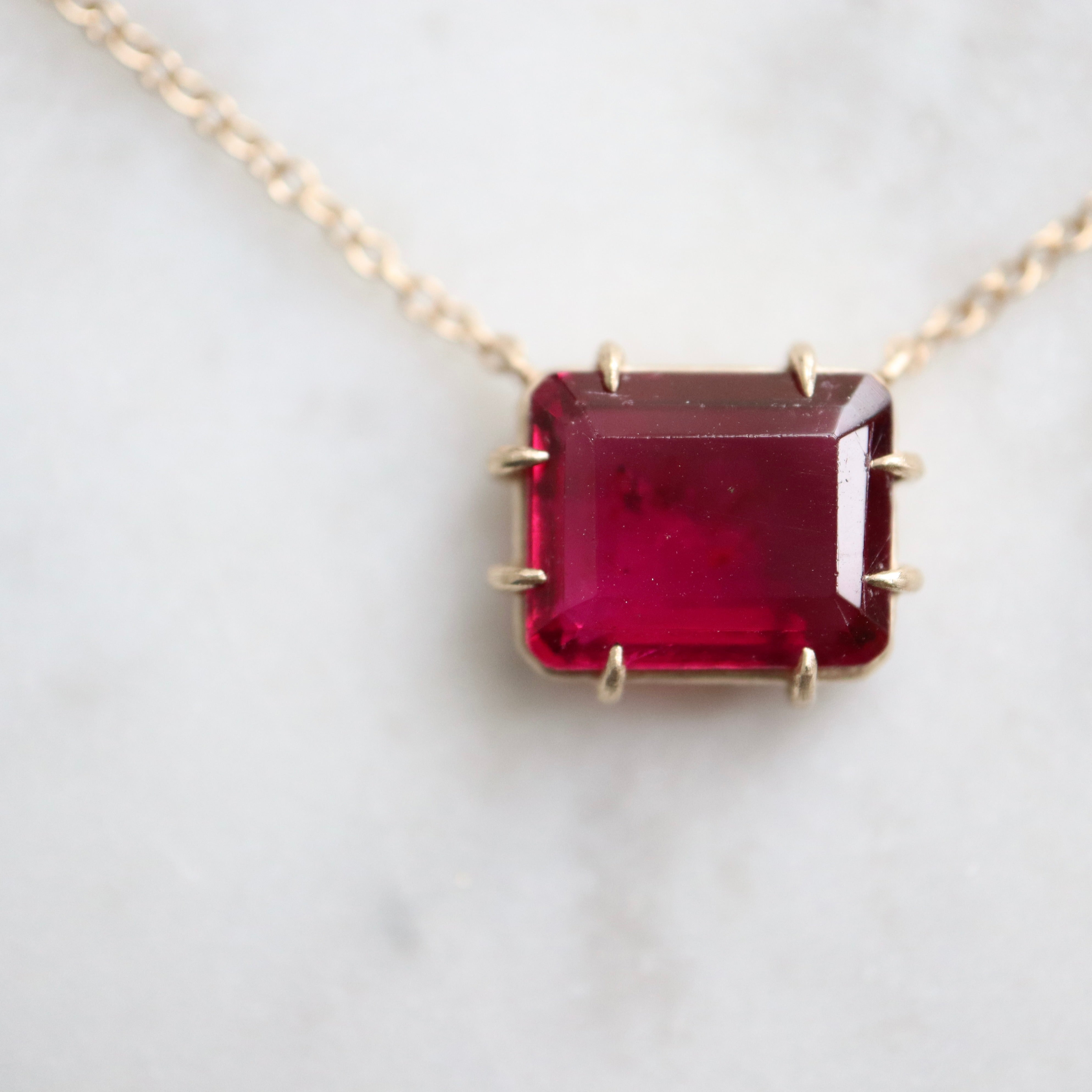 pink rectangular stone with in gold prongs on a gold chain with diamonds set in a hidden halo on the side of the setting. photographed on white marble.
