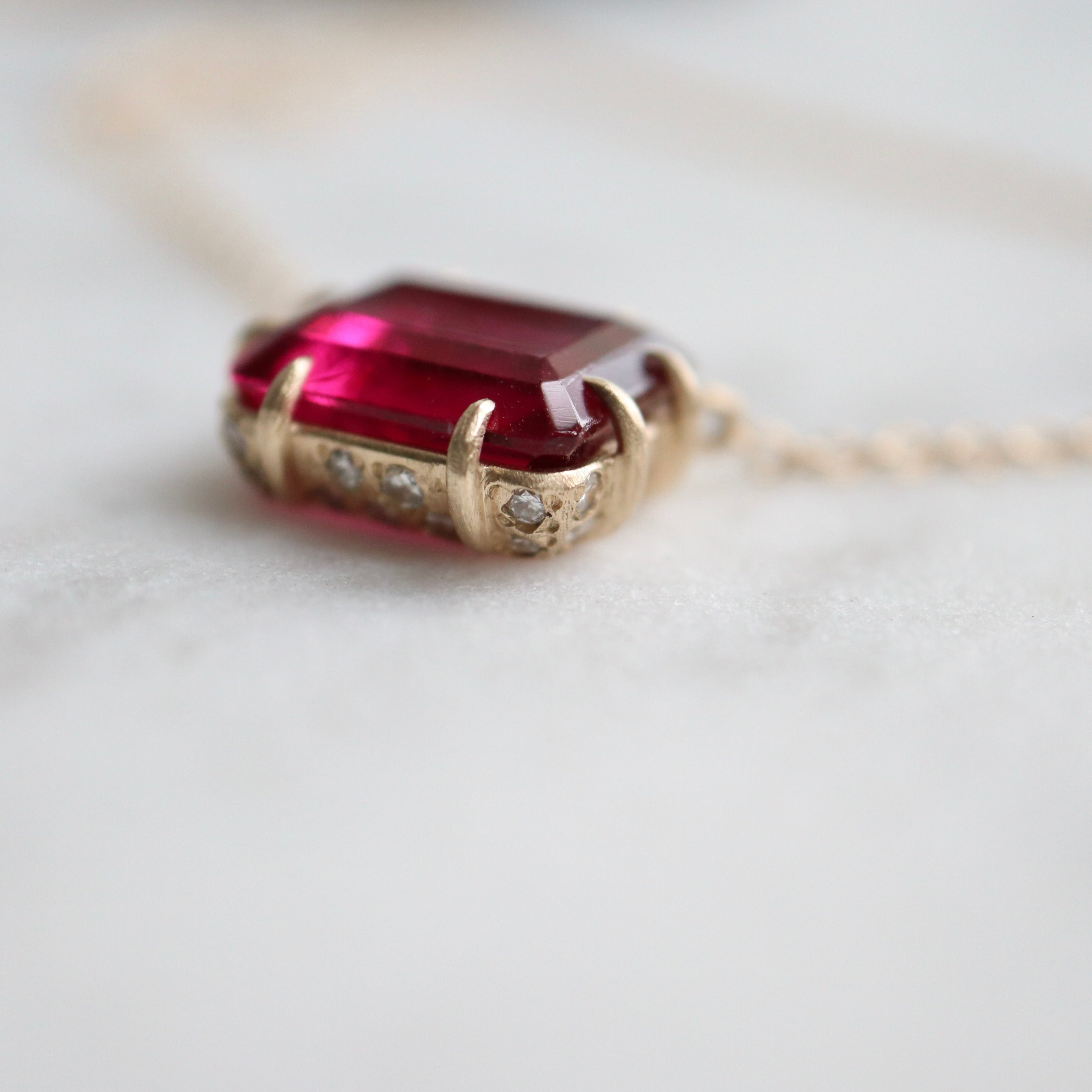 pink rectangular stone with in gold prongs on a gold chain with diamonds set in a hidden halo on the side of the setting. photographed on white marble.