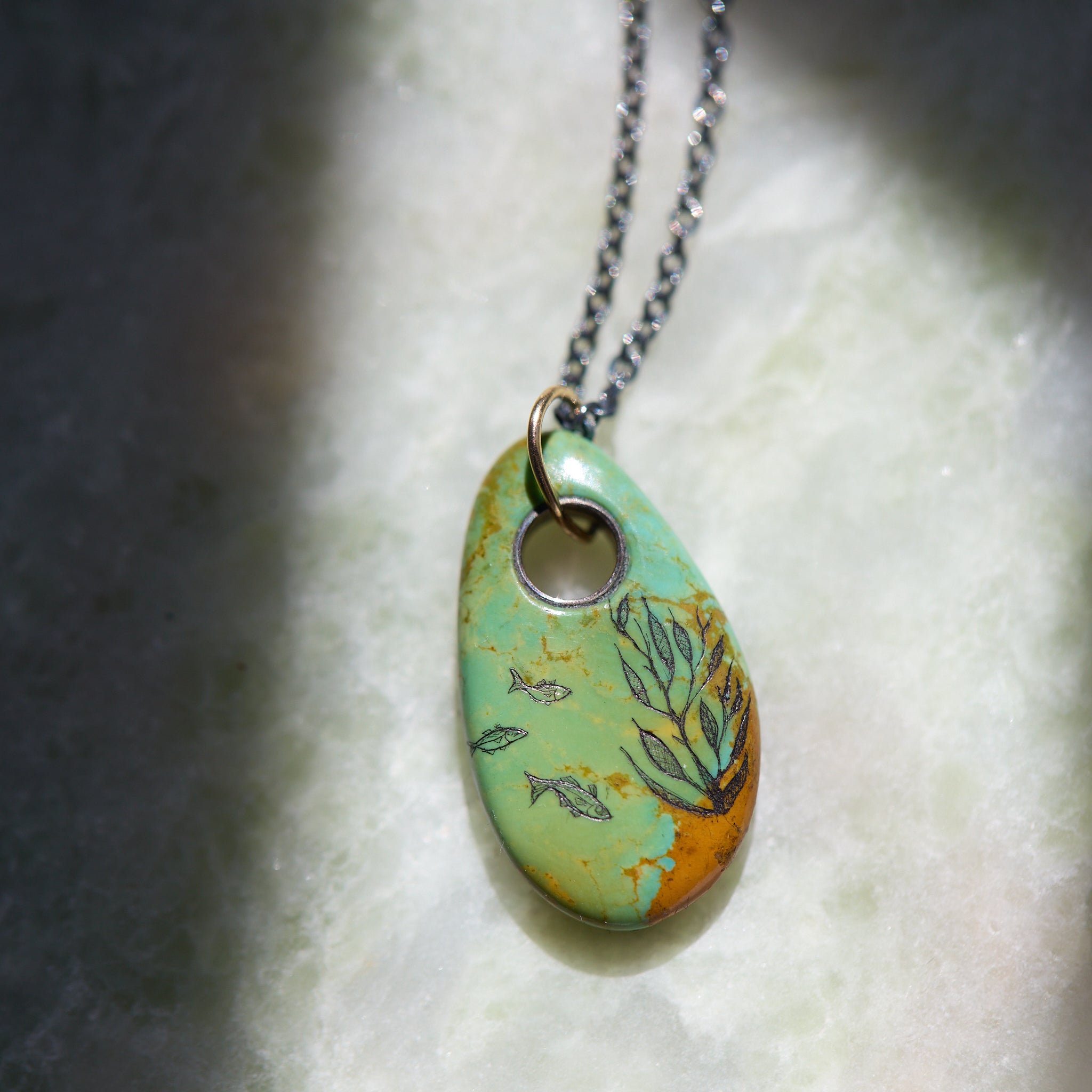 Necklace with a turquoise pendant featuring fish and seaweed illustrations on a textured surface