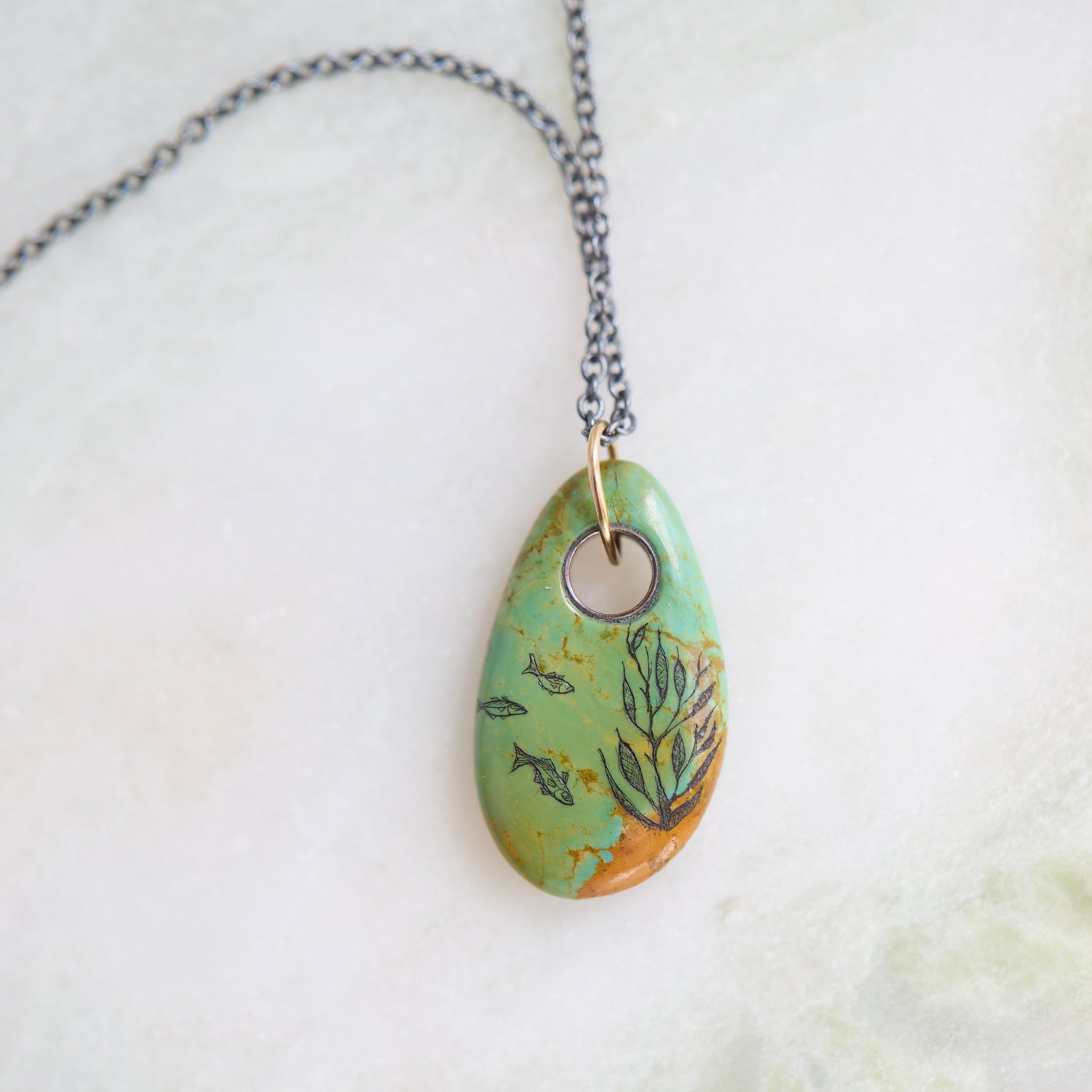 Necklace with a turquoise pendant featuring fish and seaweed illustrations on a textured surface