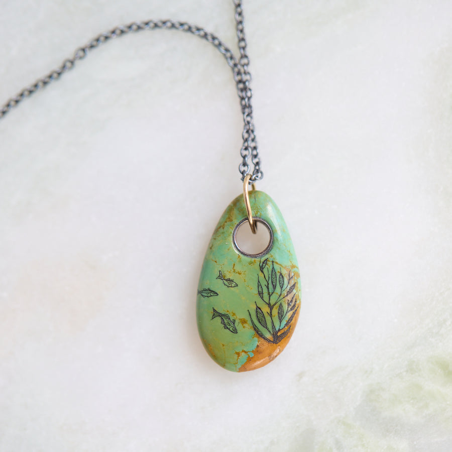 Necklace with a turquoise pendant featuring fish and seaweed illustrations on a textured surface