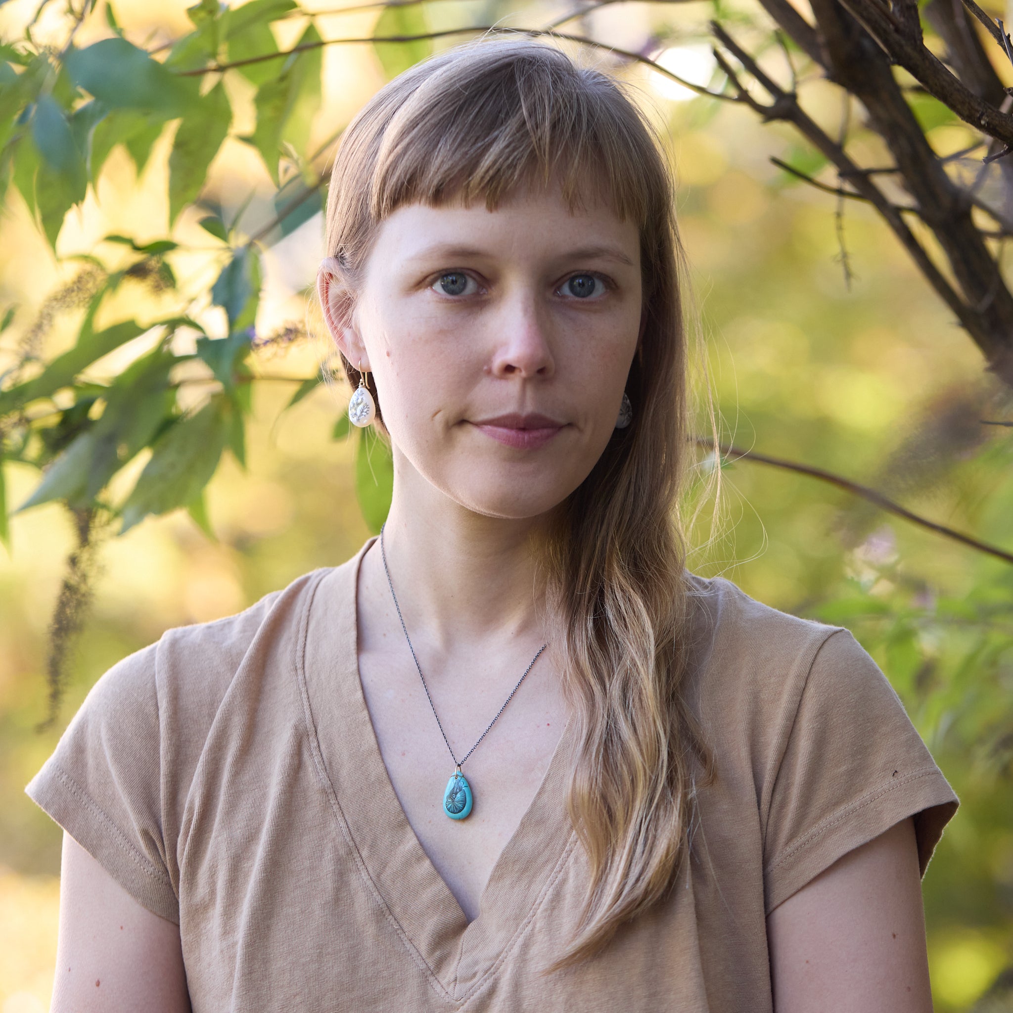 Woman with a turquoise necklace and white earrings standing in front of green foliage