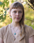Woman with a turquoise necklace and white earrings standing in front of green foliage