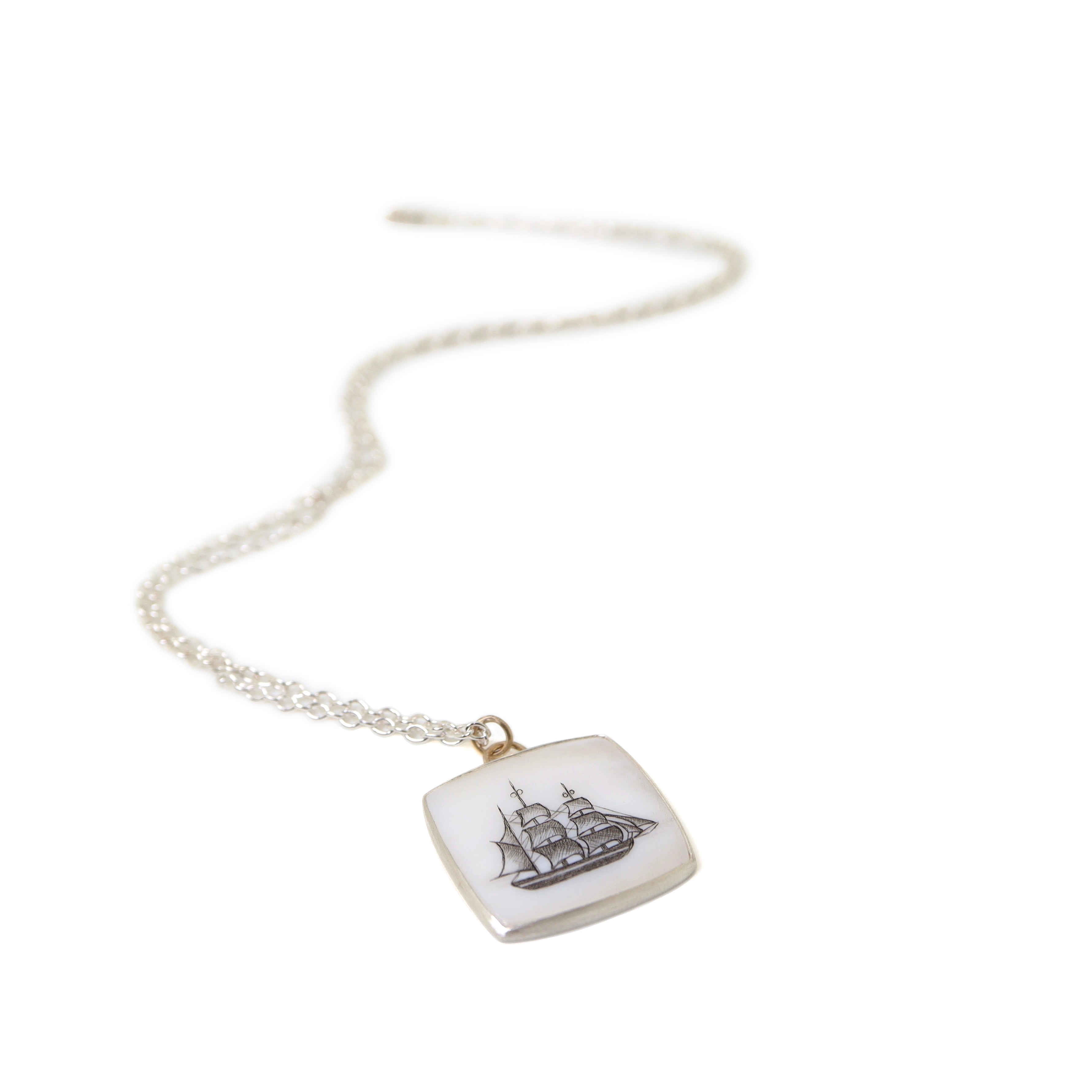 a square mother of pearl slice is set in silver with a gold ring and silver chain. An old whaling ship is hand etched into the surface with black ink, called scrimshaw.