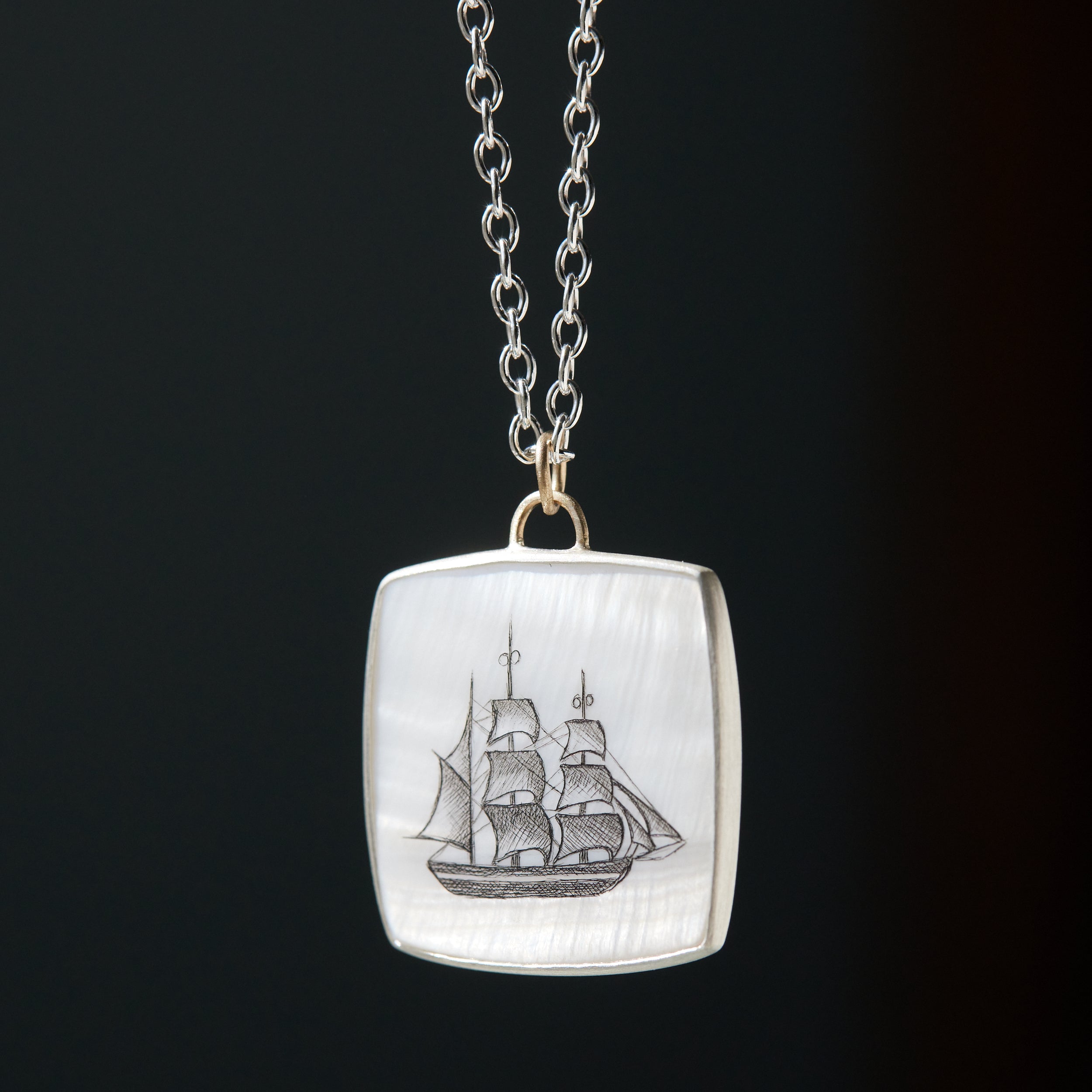 a square mother of pearl slice is set in silver with a gold ring and silver chain. An old whaling ship is hand etched into the surface with black ink, called scrimshaw.
