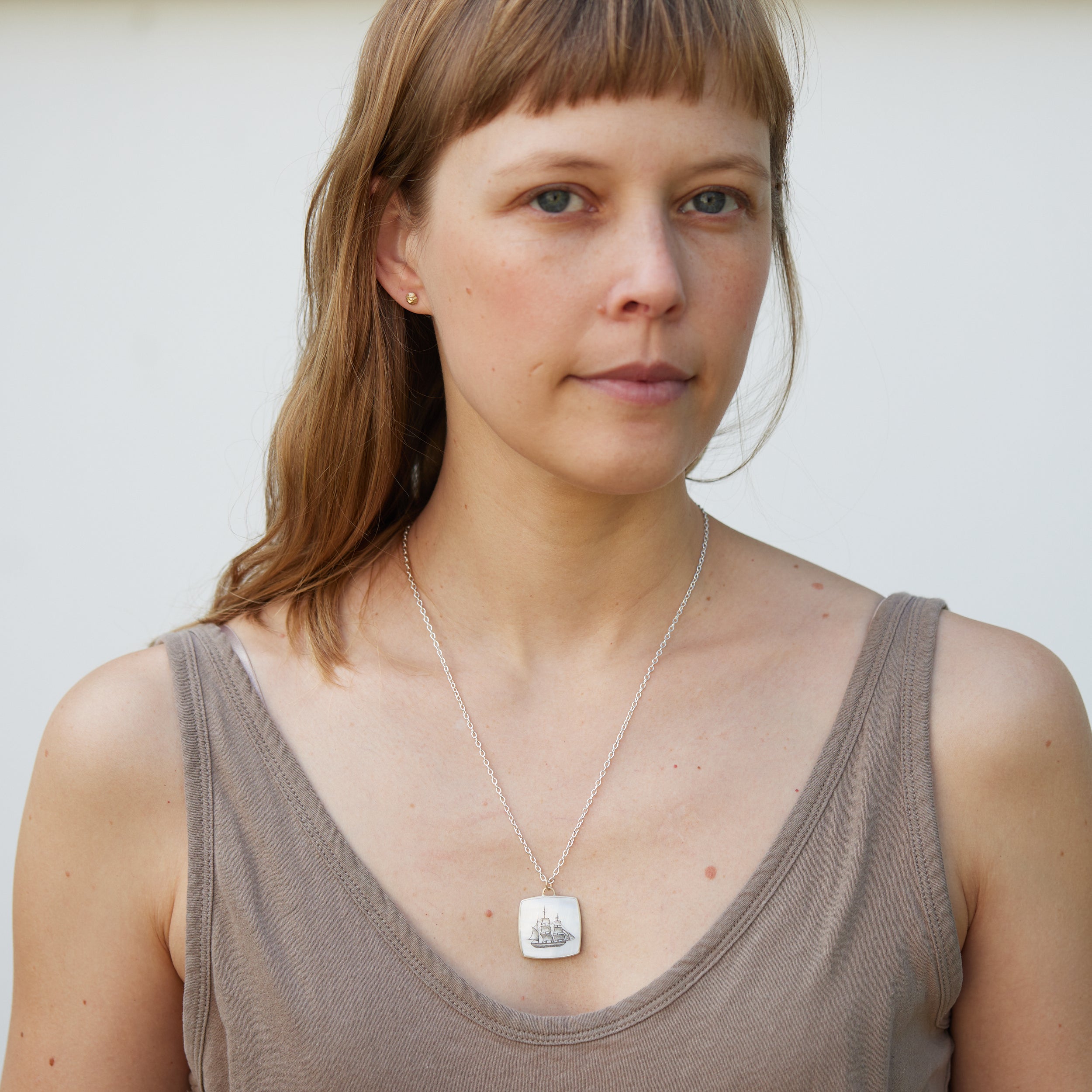 a square mother of pearl slice is set in silver with a gold ring and silver chain. An old whaling ship is hand etched into the surface with black ink, called scrimshaw.